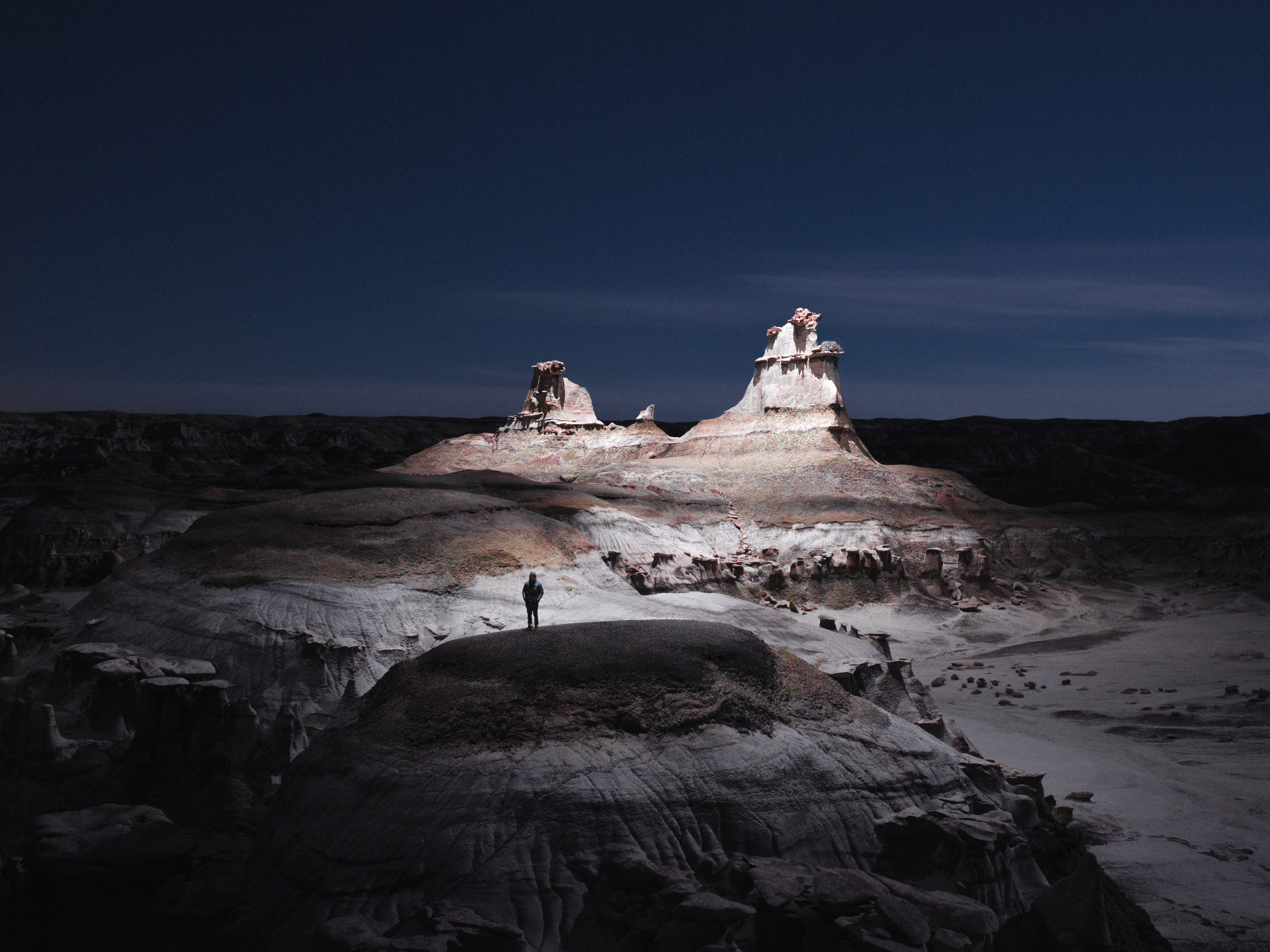A photographer stands on a rocky hill under a dark sky, with illuminated rock formations in the distance, capturing mesmerizing drone light painting displays.