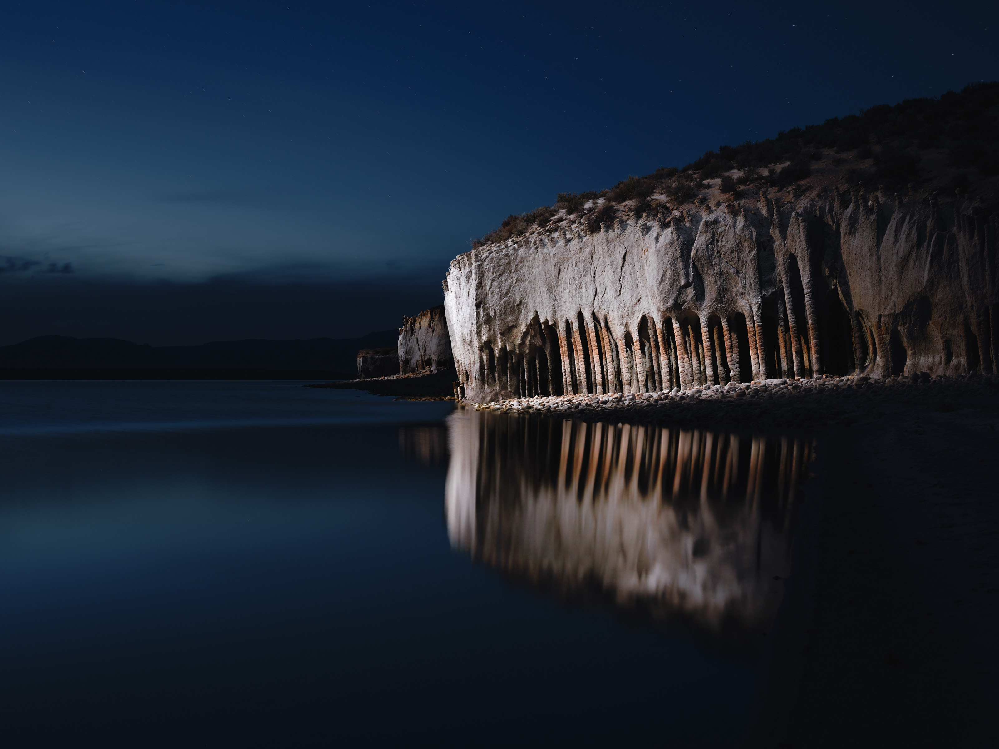 Cliff with column-like formations reflected in calm water under a dark blue night sky, subtly illuminated by ethereal patterns of drone light art.