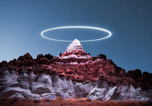 A glowing white ring hovers above a rocky mountain peak under a starry night sky, creating a mesmerizing scene of drone light painting in this nighttime landscape photography.