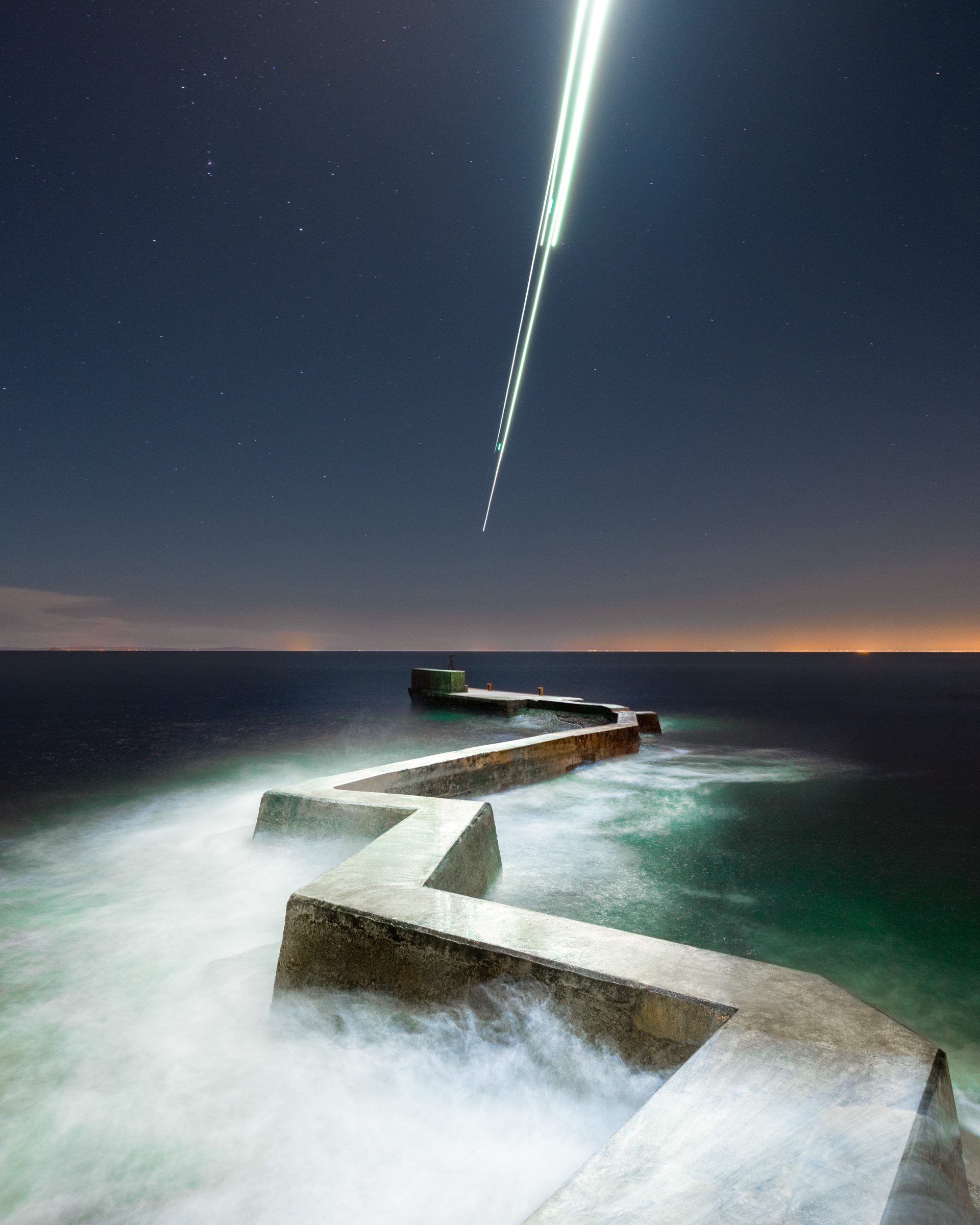 A zigzag sea wall extends into the ocean under a starry night sky, with mesmerizing drone light painting creating bright trails above, capturing the essence of nighttime landscape photography.