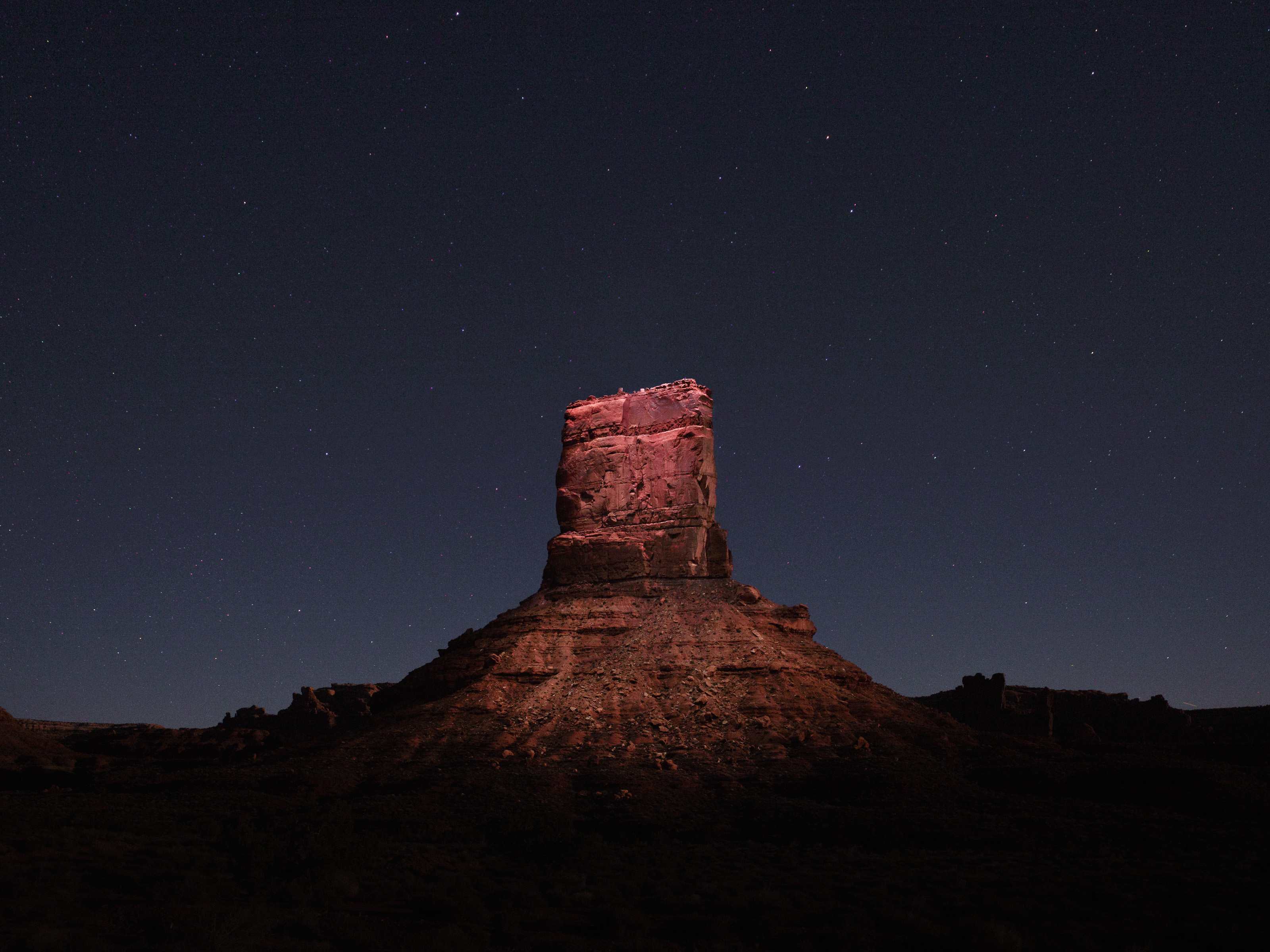 A rocky butte at night, illuminated by futuristic pink drone light art under a starry sky.