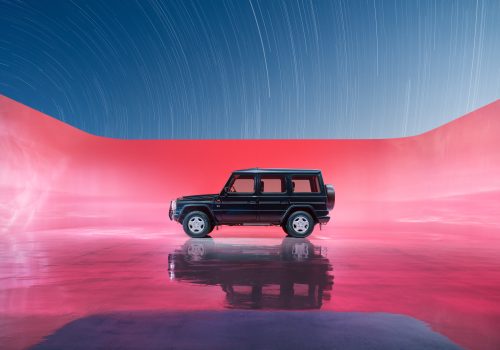 A black SUV on a reflective pink surface with a pink wall and star trails—enhanced by futuristic drone light painting dancing in the night sky above.