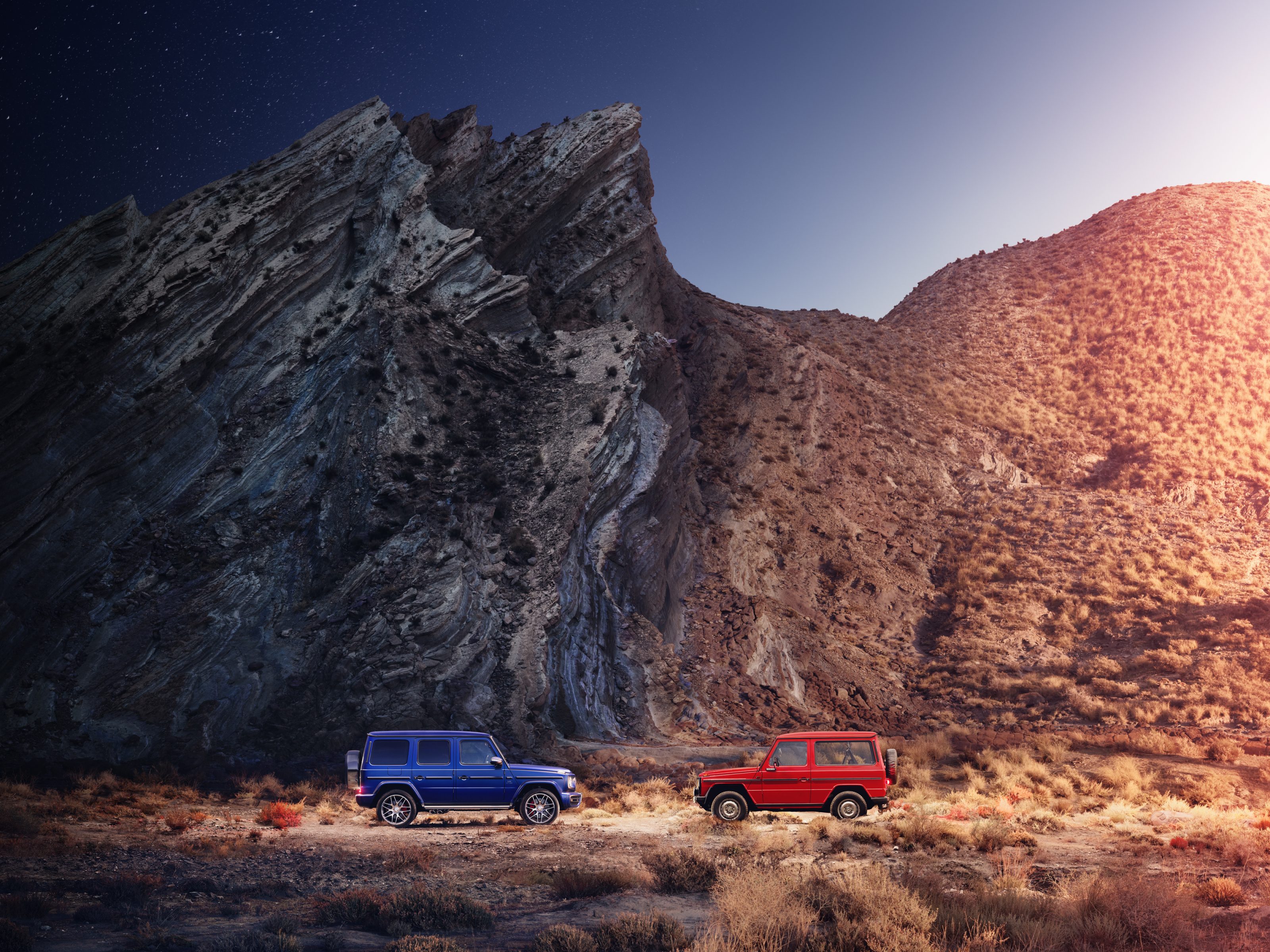 Two SUVs, one blue and one red, parked on rocky terrain beside a jagged mountain under a clear sky, as a photographer prepares to capture the dramatic landscape.