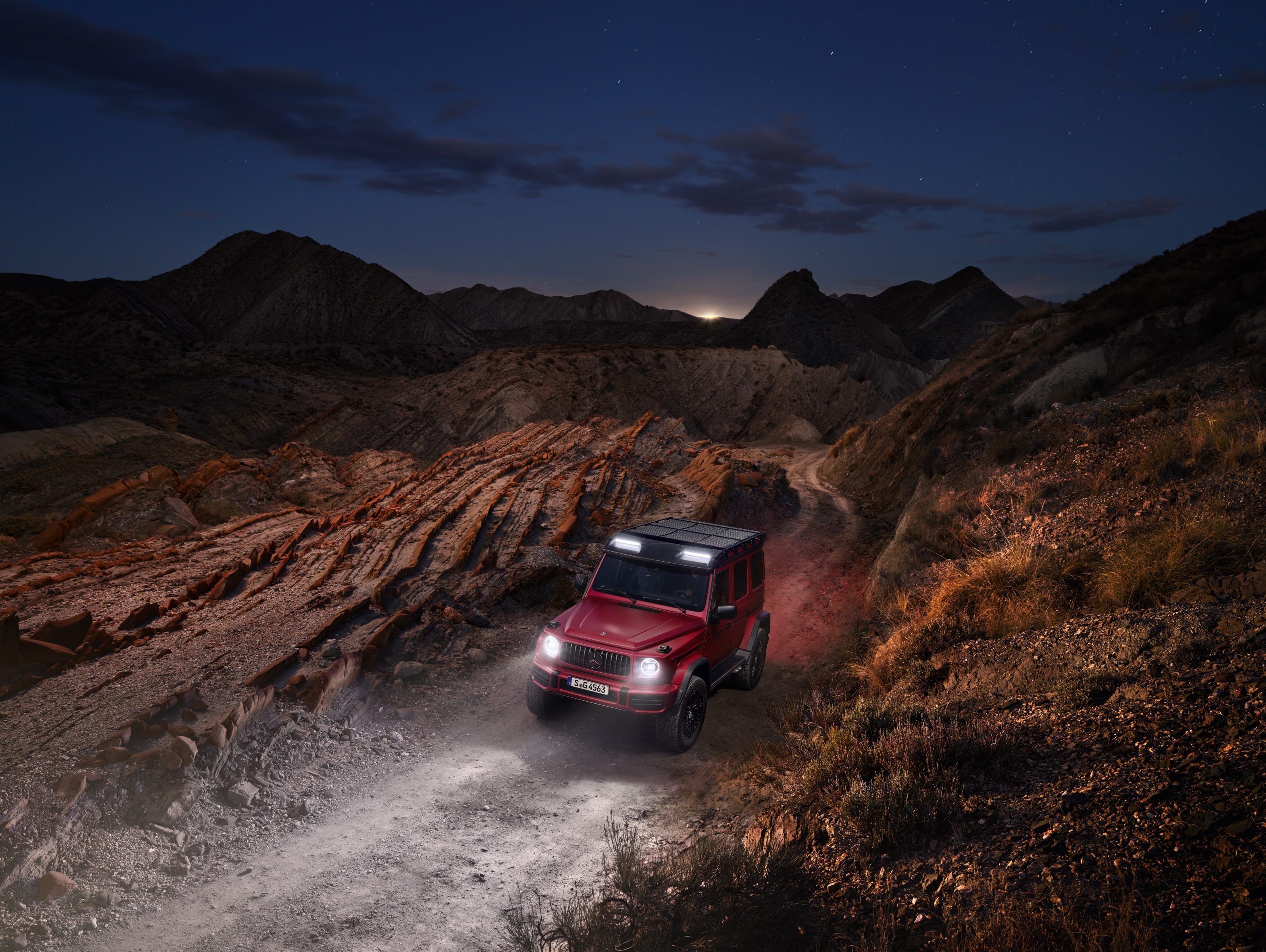A red off-road SUV drives on a dusty trail through rocky hills at dusk under a twilight sky, illuminated by futuristic drone light art swirling above.