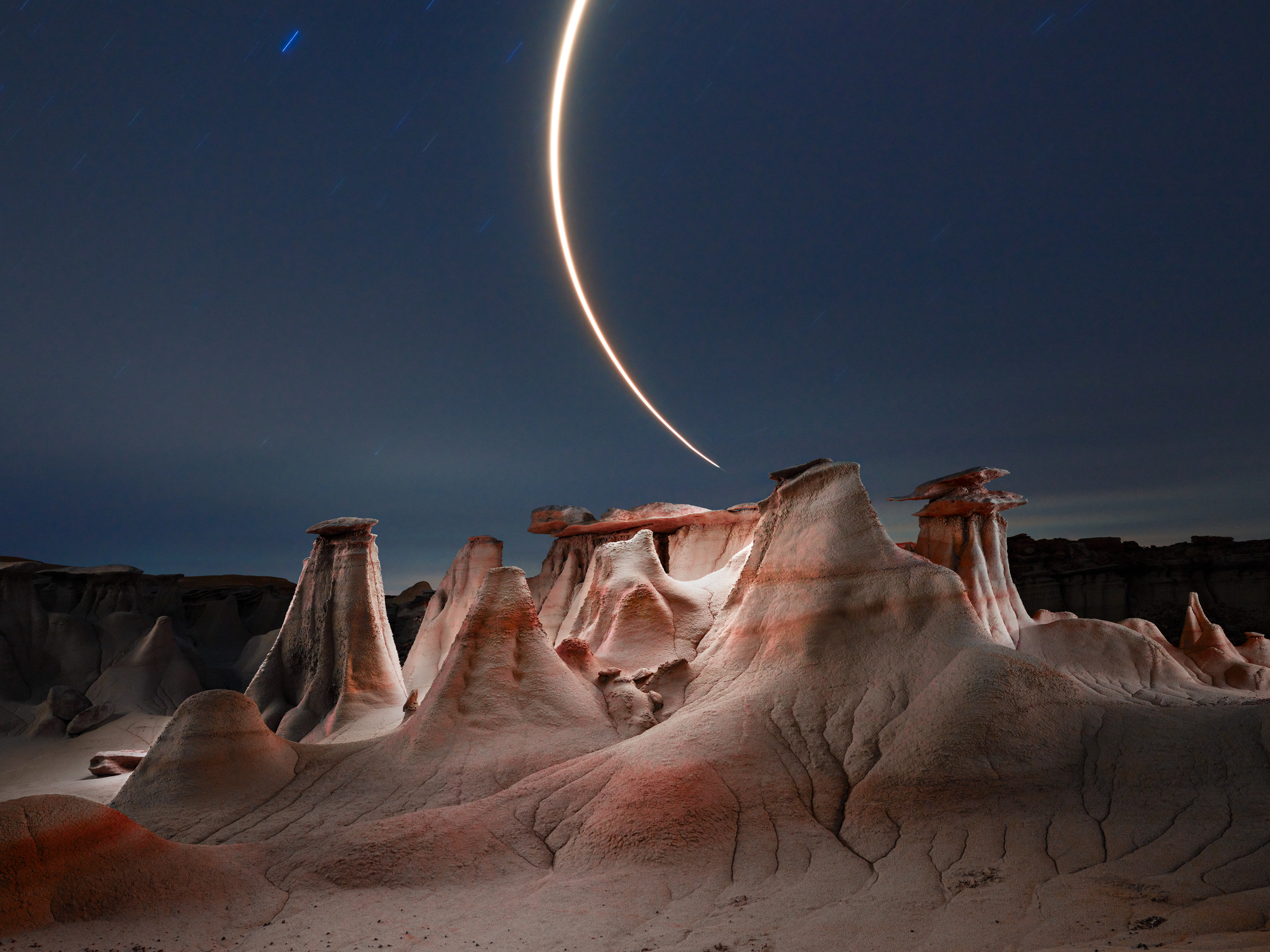 Eroded rock formations at night with a bright crescent moon trail arcing across the sky, enhanced by mesmerizing drone light painting that adds an ethereal touch to the landscape.