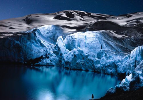A person stands by a glowing blue icy lake beneath snowy mountains, as futuristic drone light art illuminates the starry night sky.