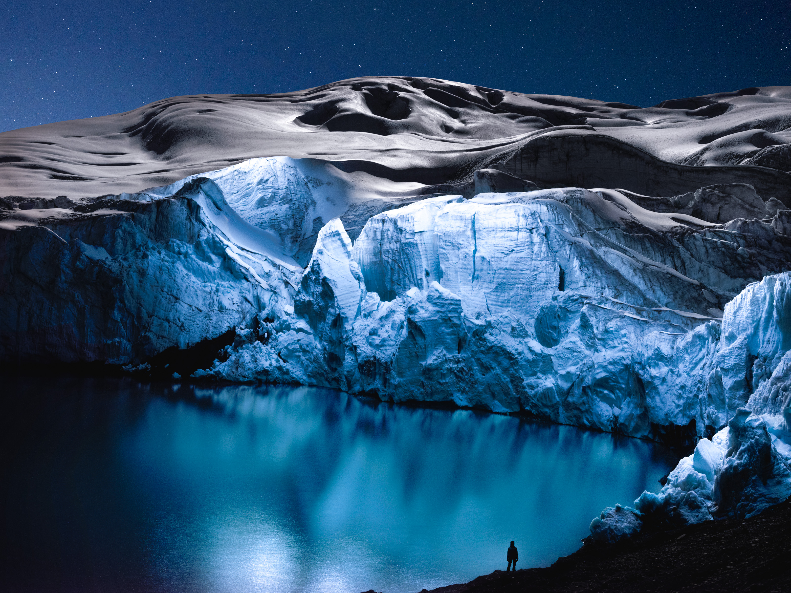 A person stands by a glowing blue icy lake beneath snowy mountains, as futuristic drone light art illuminates the starry night sky.