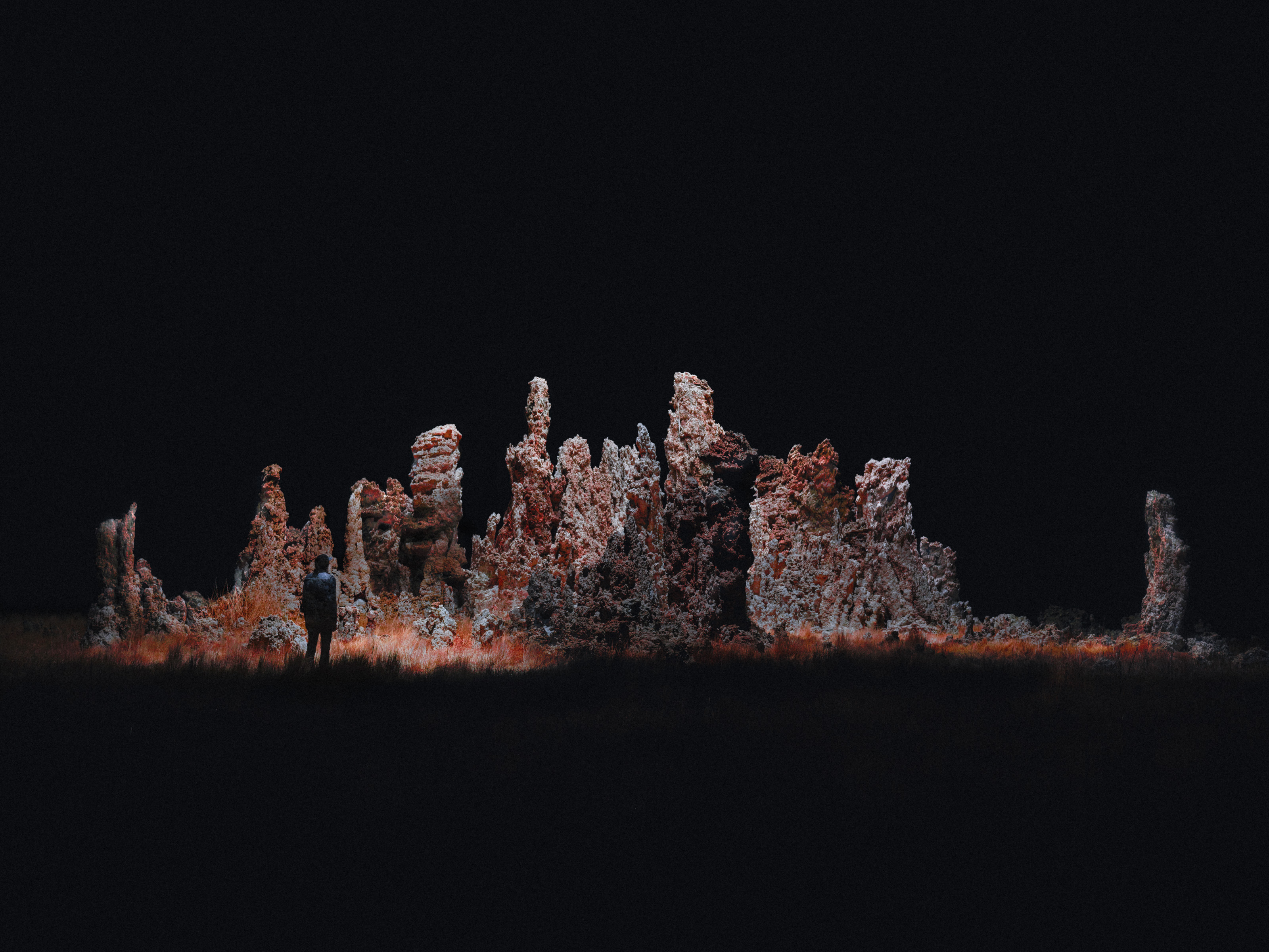 A person stands near tall, jagged rock formations illuminated in the darkness, as futuristic drone light art adds a surreal glow to the nighttime landscape.