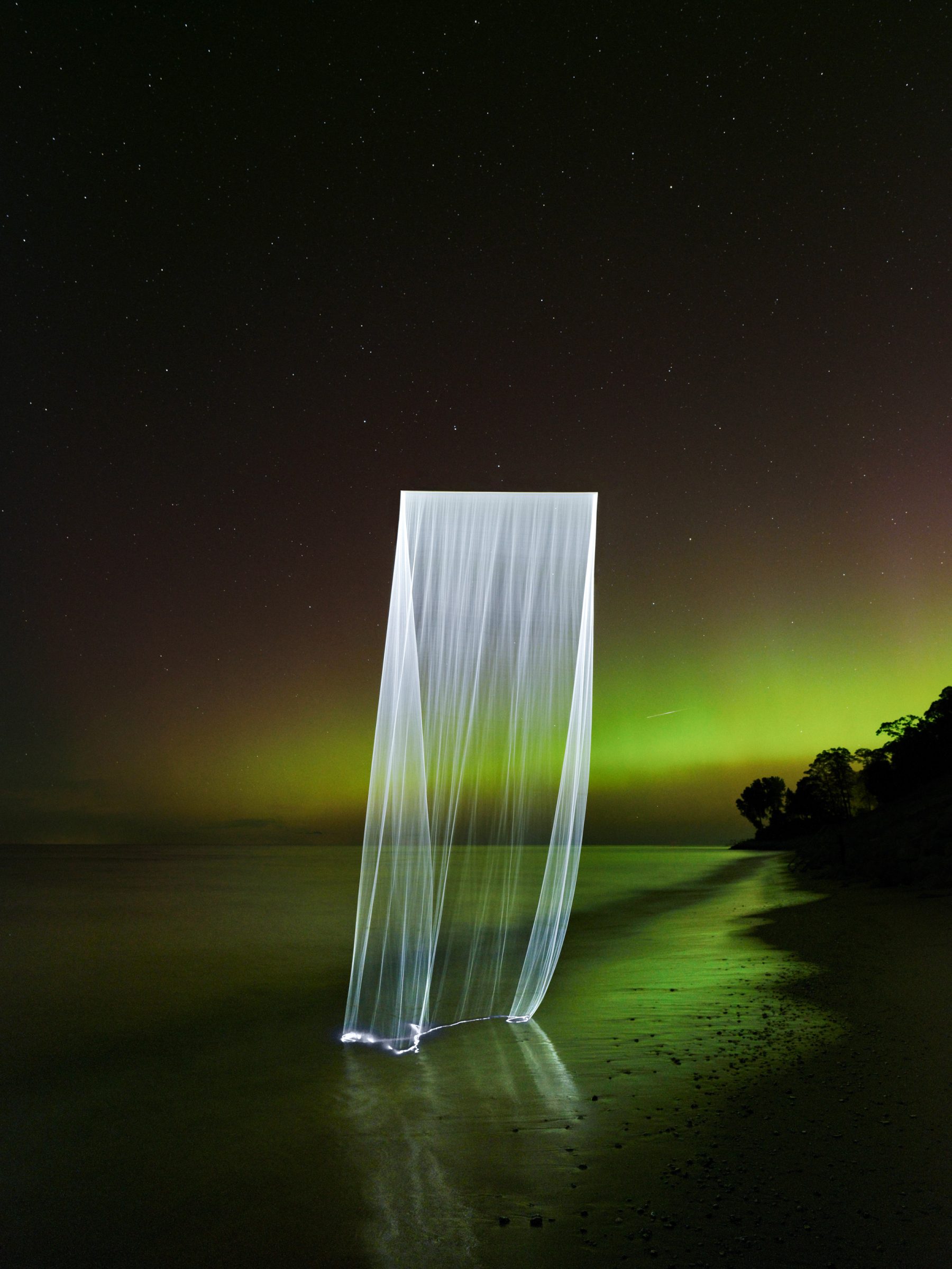 A glowing, translucent curtain-like light stands on a beach at night, with green aurora and stars above. This Siren Series Photograph captures drone light art in a mesmerizing display of a fine artist’s vision.