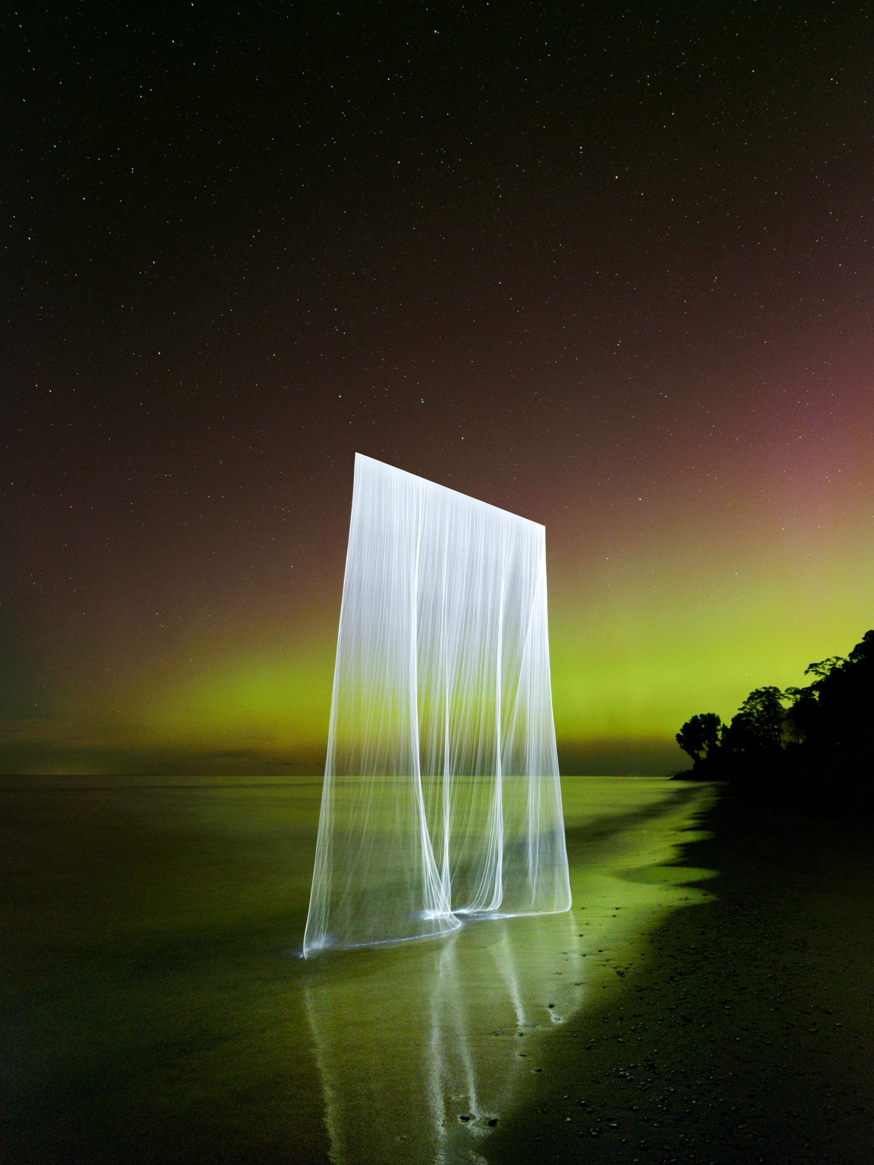 A glowing, translucent sheet of light—crafted through drone light painting—stands on a beach under a starry sky with green aurora in the background, capturing the imagination of every photographer and fine artist.