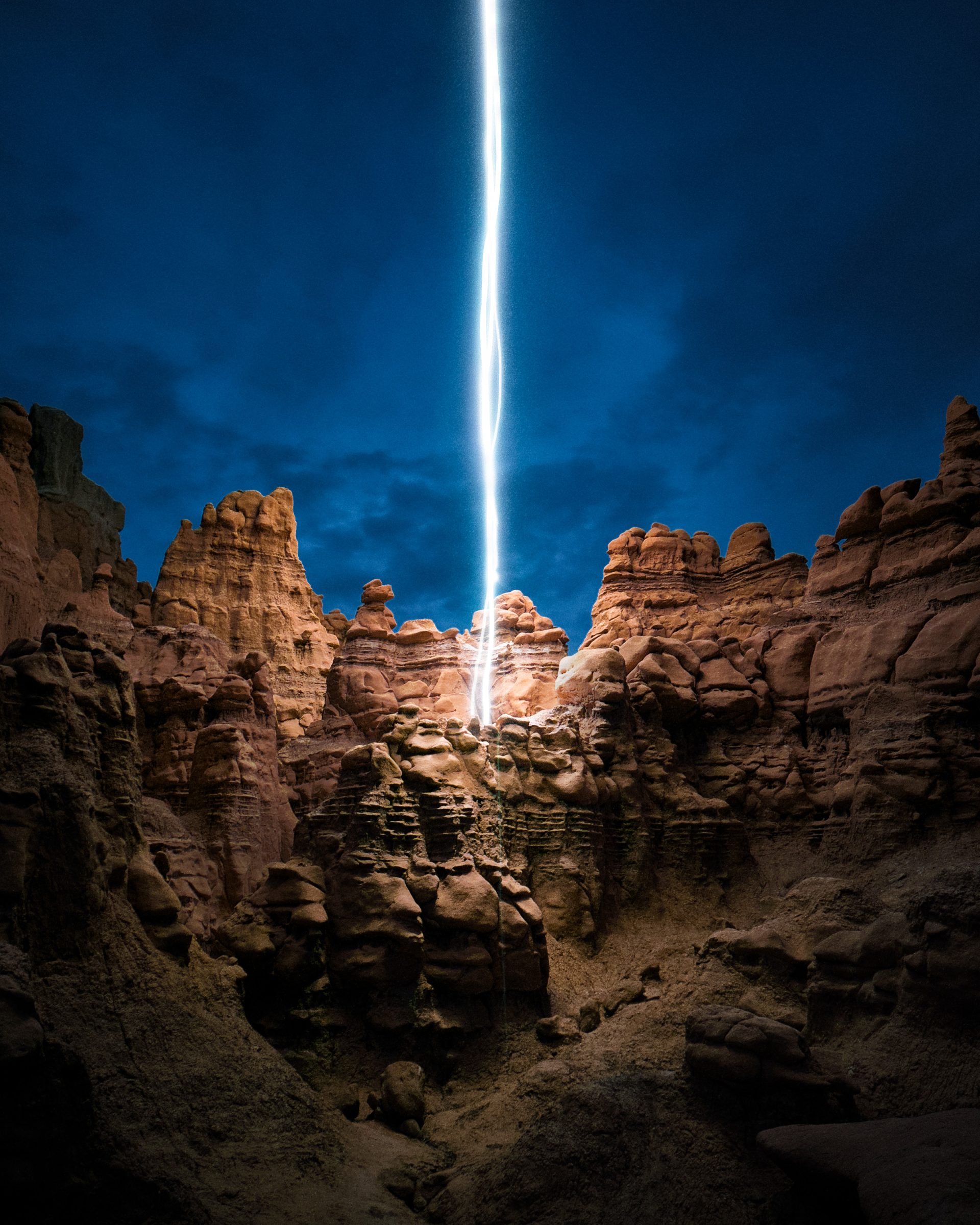 A bolt of lightning strikes red rock formations under a dark, cloudy blue sky at night, capturing the drama of nighttime landscape photography.