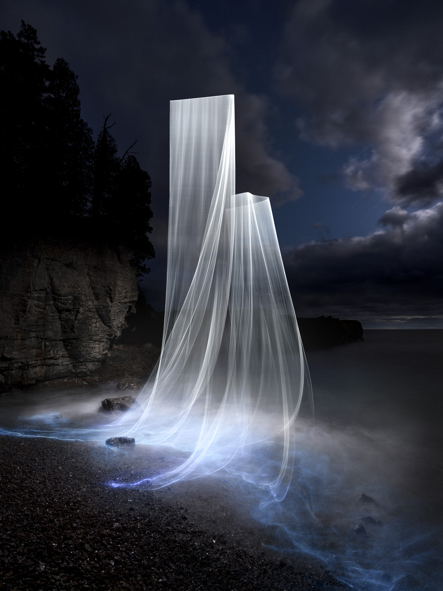 Tall, glowing white light trails rise from a rocky shore at night in this captivating Siren Series nighttime landscape photography, with dramatic cliffs and clouds forming a stunning backdrop.