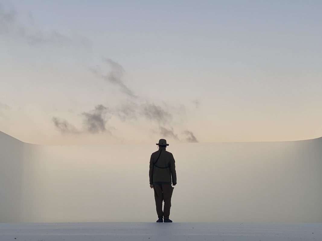 A photographer in a hat stands alone, facing a minimalist, curved wall under a soft, cloudy sky, capturing the scene’s quiet, futuristic atmosphere.