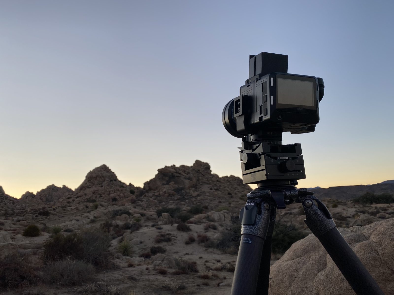 A photographer has set up a camera on a tripod in a rocky desert landscape at dusk, capturing the beauty of nighttime landscape photography.