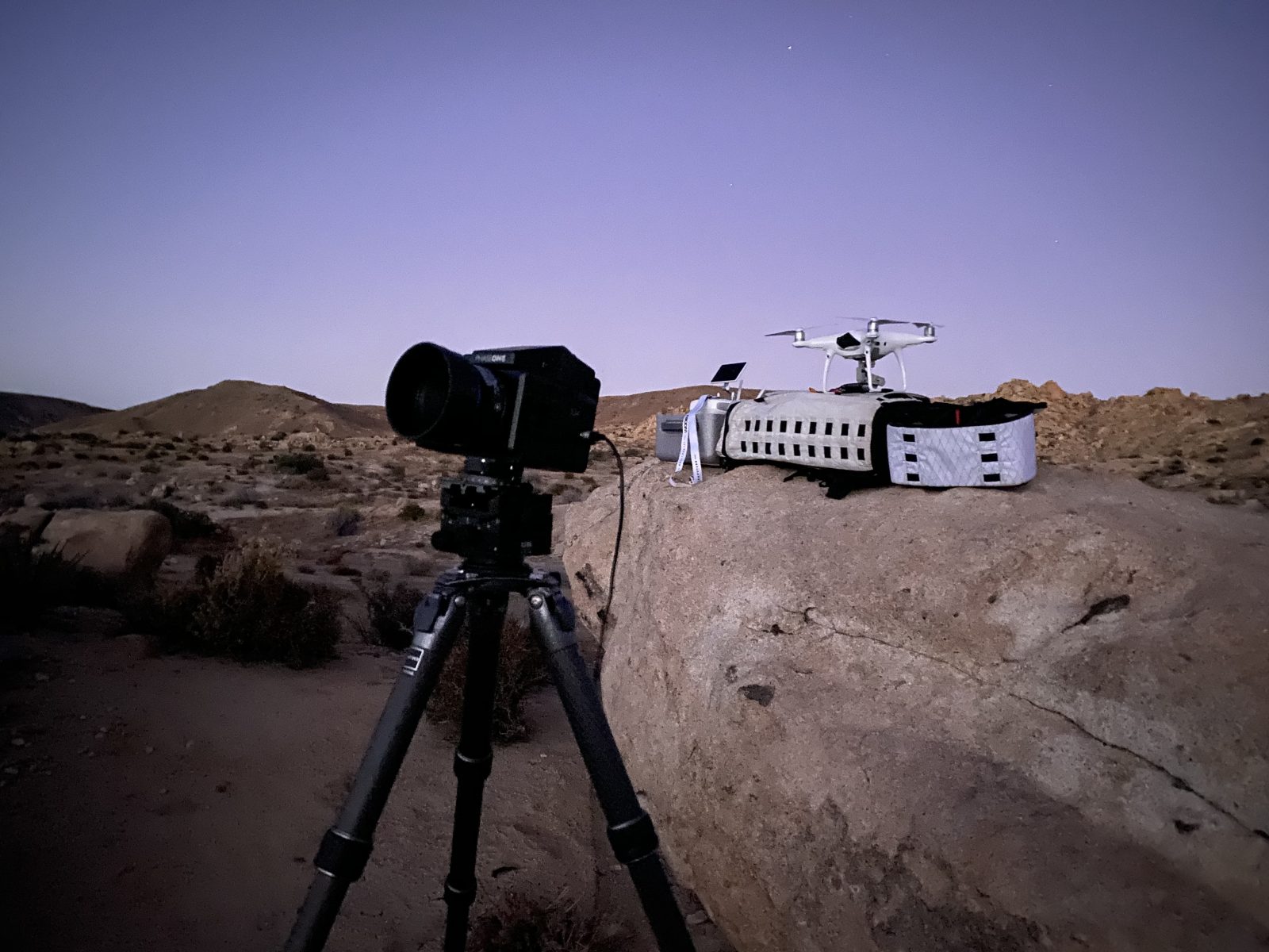 A photographer captures a futuristic scene with a camera on a tripod and a drone on a rock in the desert at dusk, ready for drone light painting.