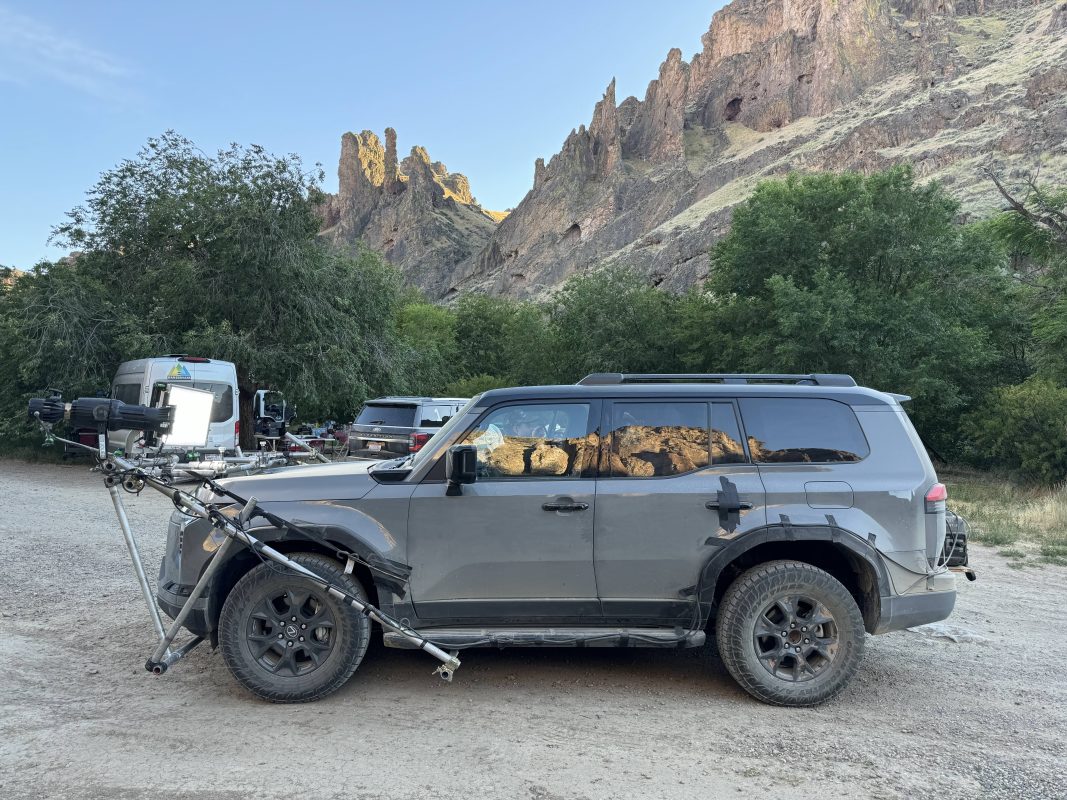 A Lexus GX550 SUV equipped with a camera rig parked on a gravel surface, surrounded by lush vegetation and rocky formations in the background.