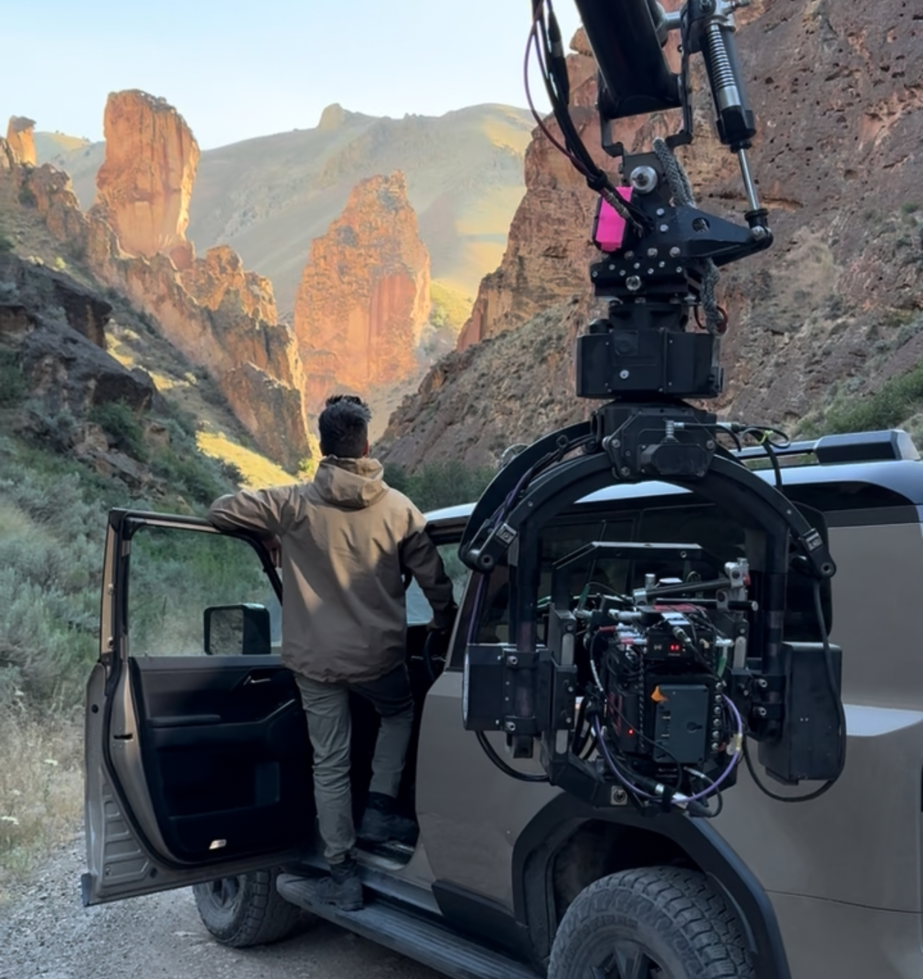 A person stands on the side of an SUV with an open door, gazing at towering rock formations in a canyon during daylight.