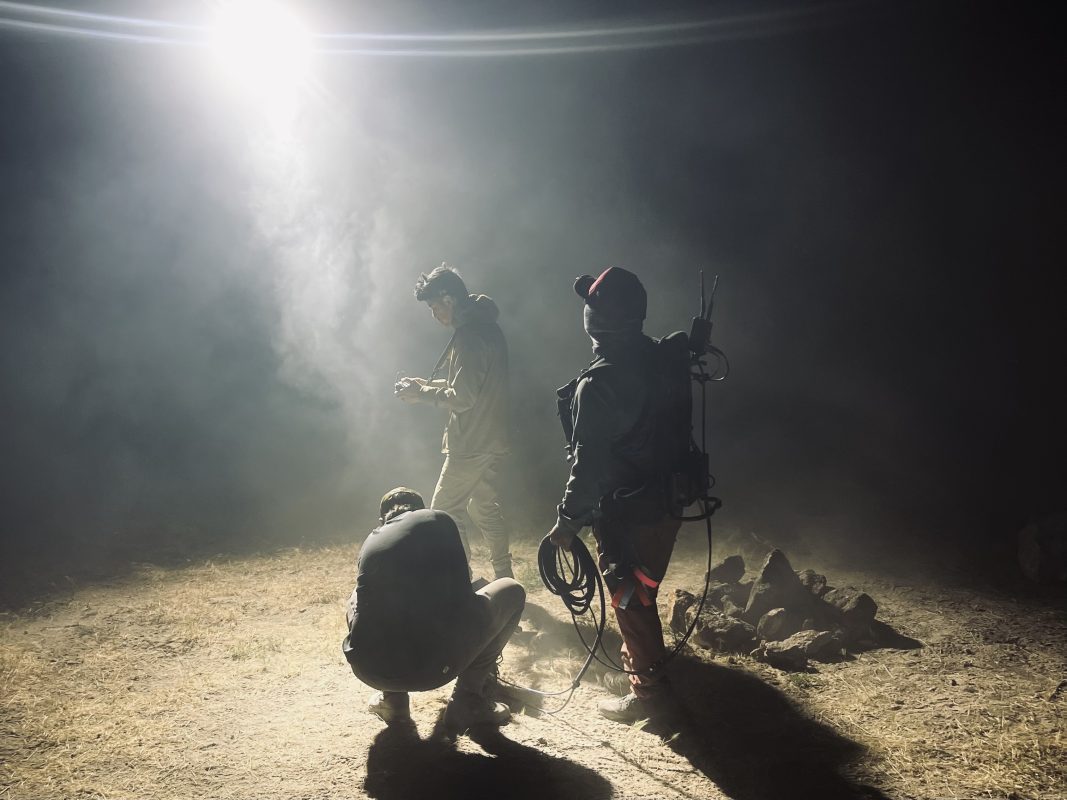 Three crew members work in a smoky environment at night, illuminated by a strong light source. One person stands while holding equipment, another crouches down, and the third looks on with a headset.