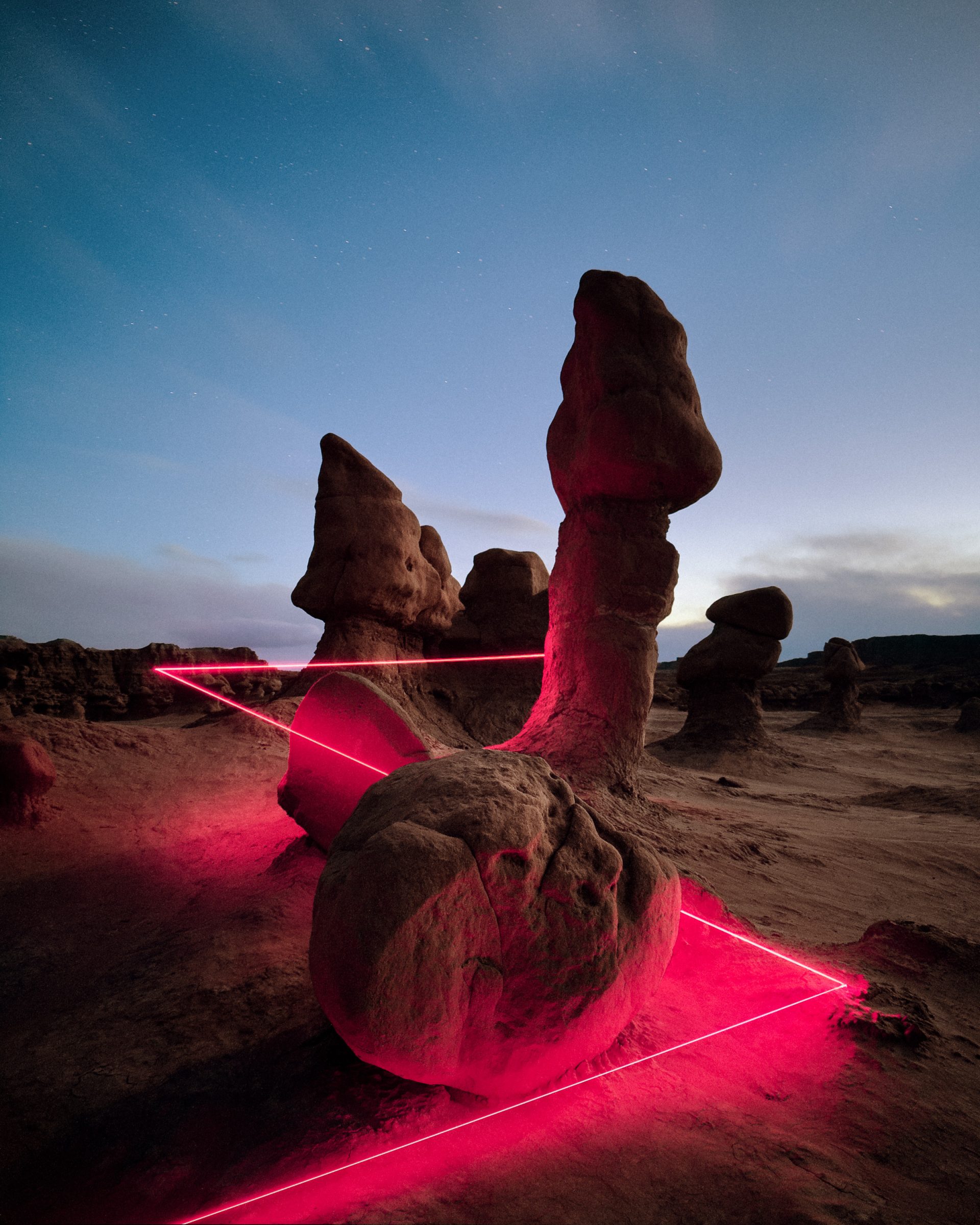 Rock formations in a desert at dusk illuminated by pink laser lights and vibrant drone light art, set against a tranquil blue sky.