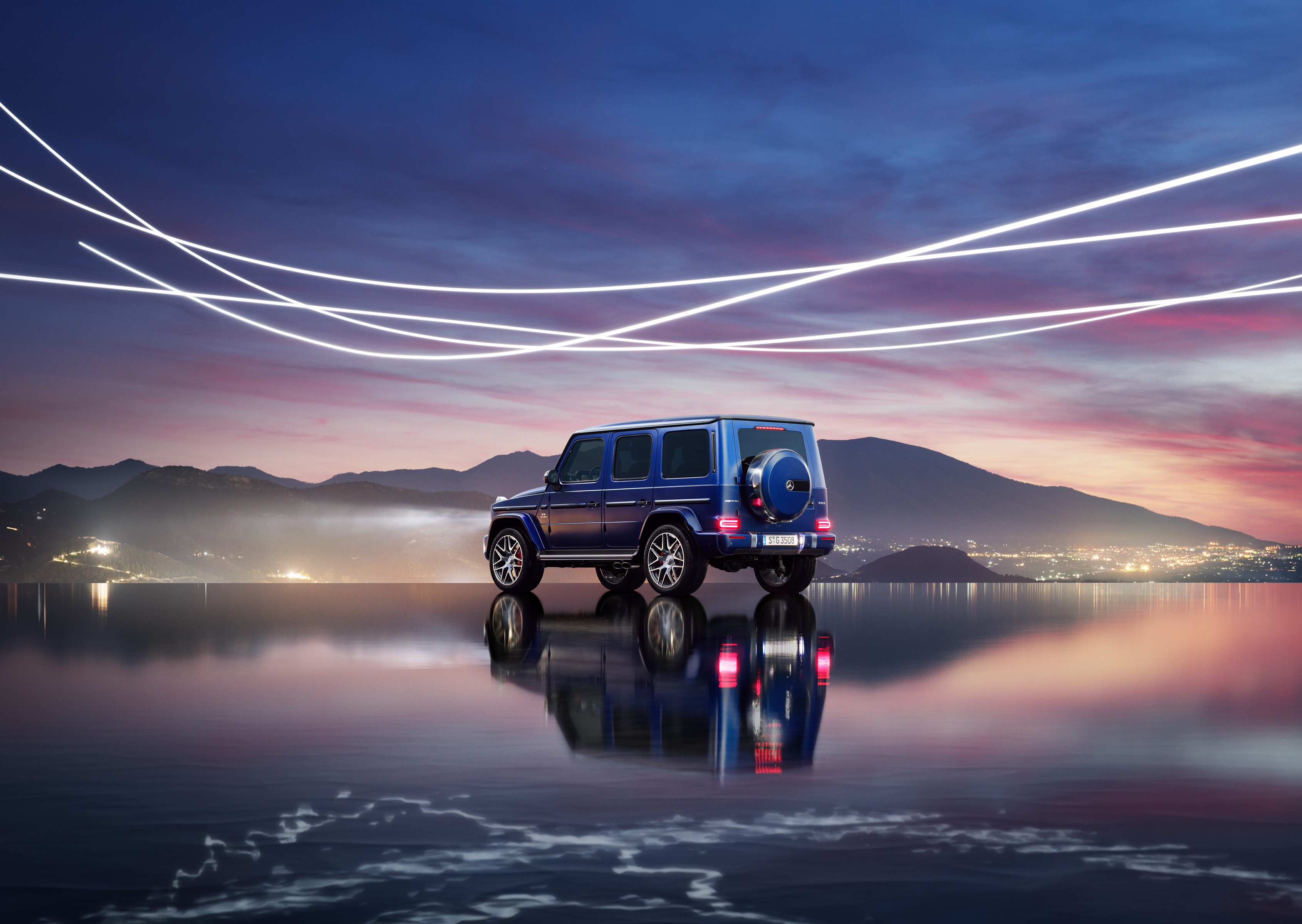 A blue SUV parked by reflective water at sunset, with drone light art creating dynamic trails in the sky and mountains in the background.