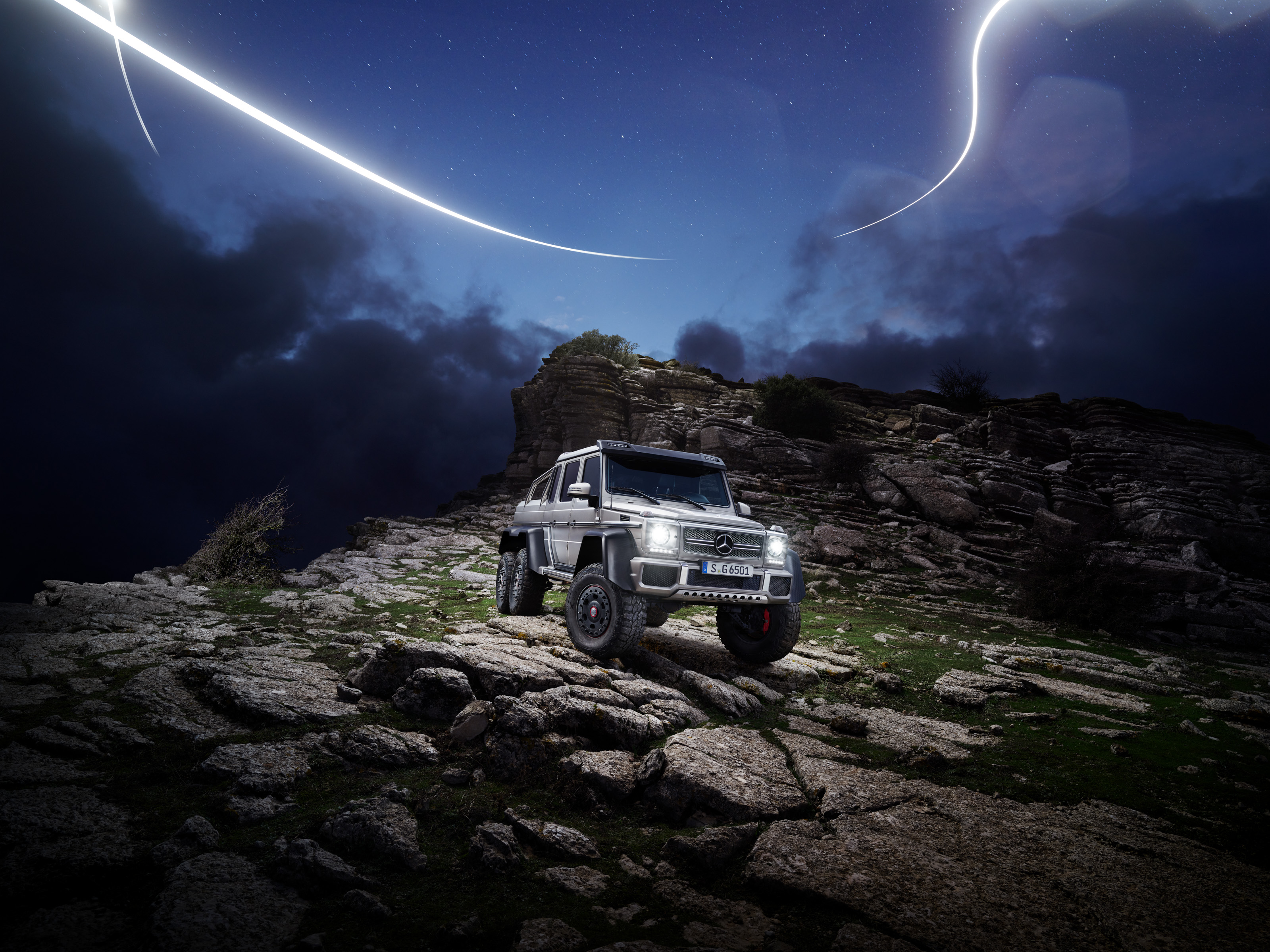 A silver SUV on rocky terrain at night, captured by a photographer, with dramatic clouds and streaks of light creating a futuristic nighttime landscape photography scene.