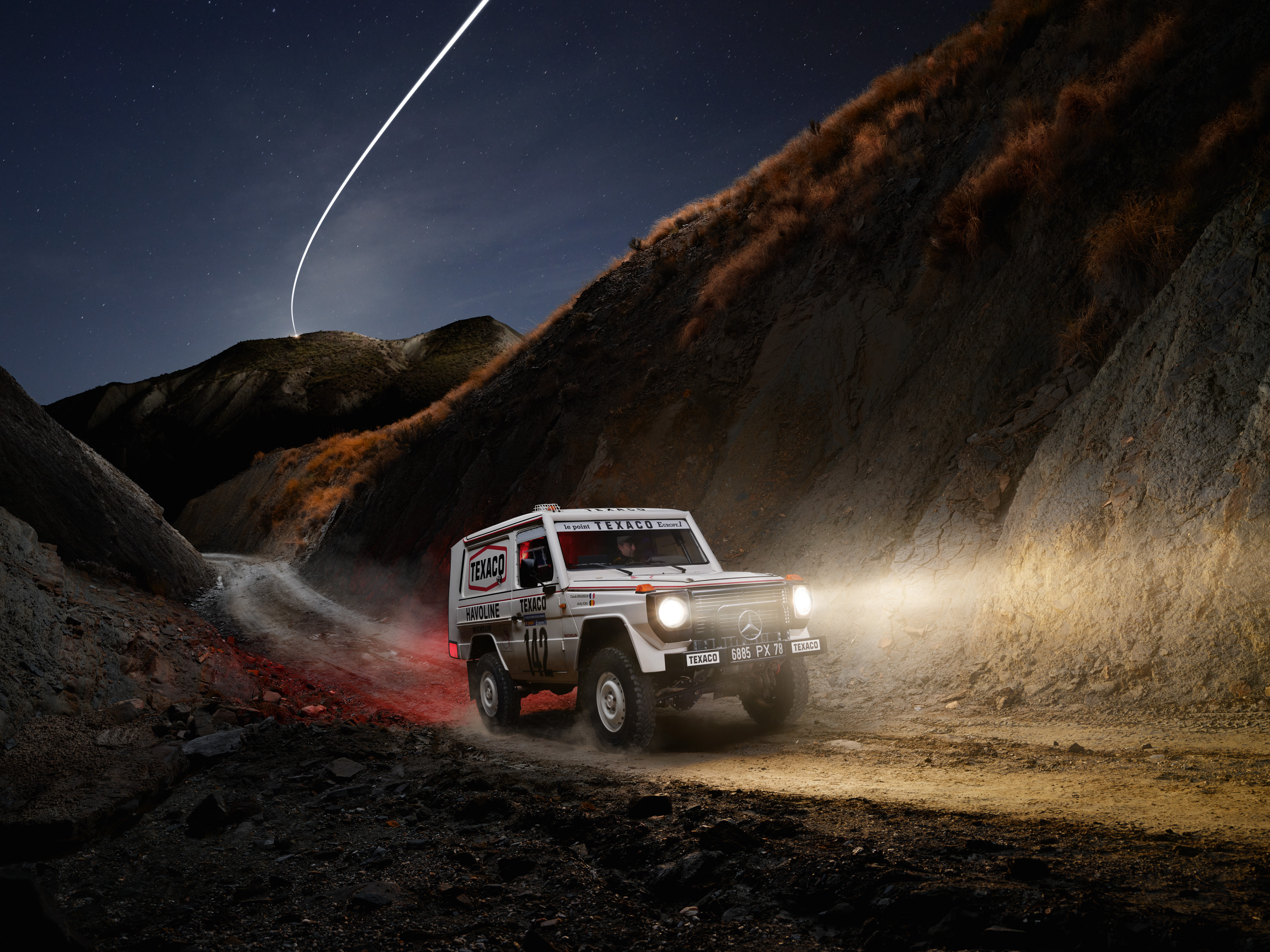 A Mercedes-Benz off-road vehicle drives on a dirt mountain trail at night, its headlights illuminating the path like a scene from nighttime landscape photography.