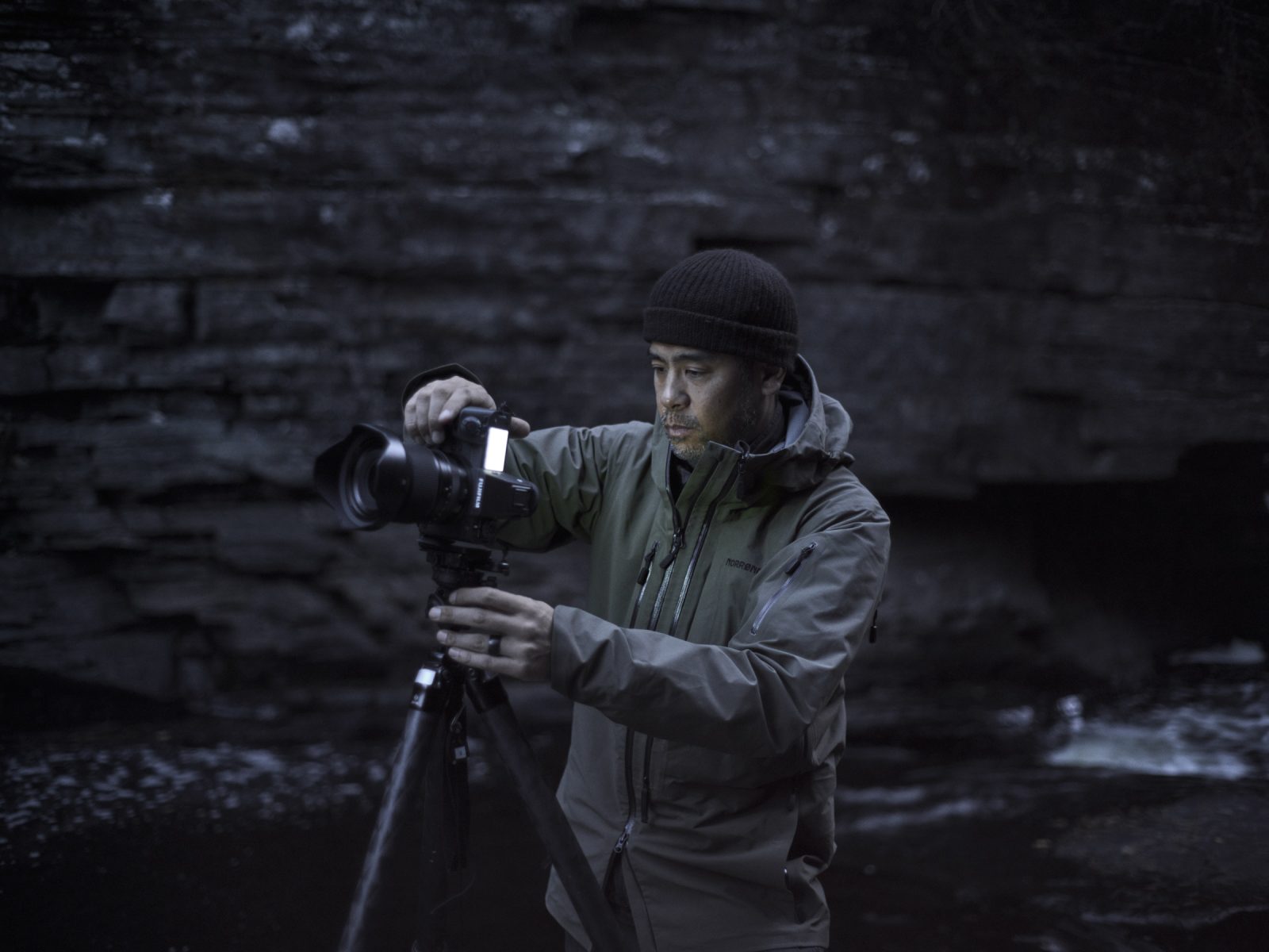 A man in a jacket adjusts a camera on a tripod by a rocky stream in low light, preparing for nighttime landscape photography.