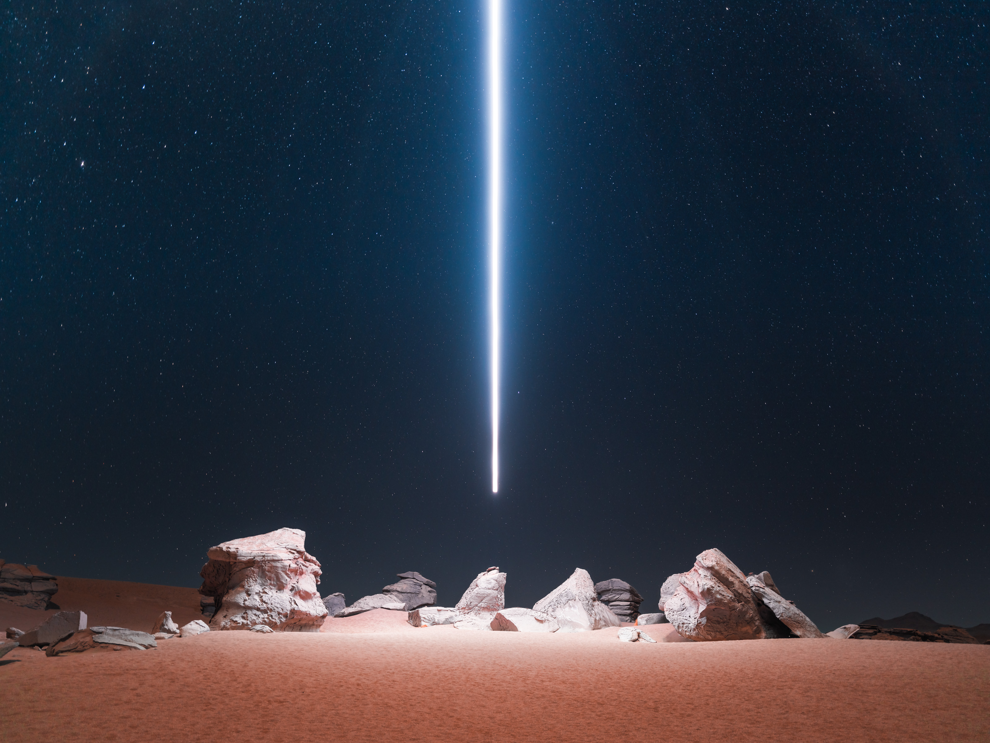 A bright beam of light, created through drone light painting, descends from the night sky onto rocks in a desert nighttime landscape.