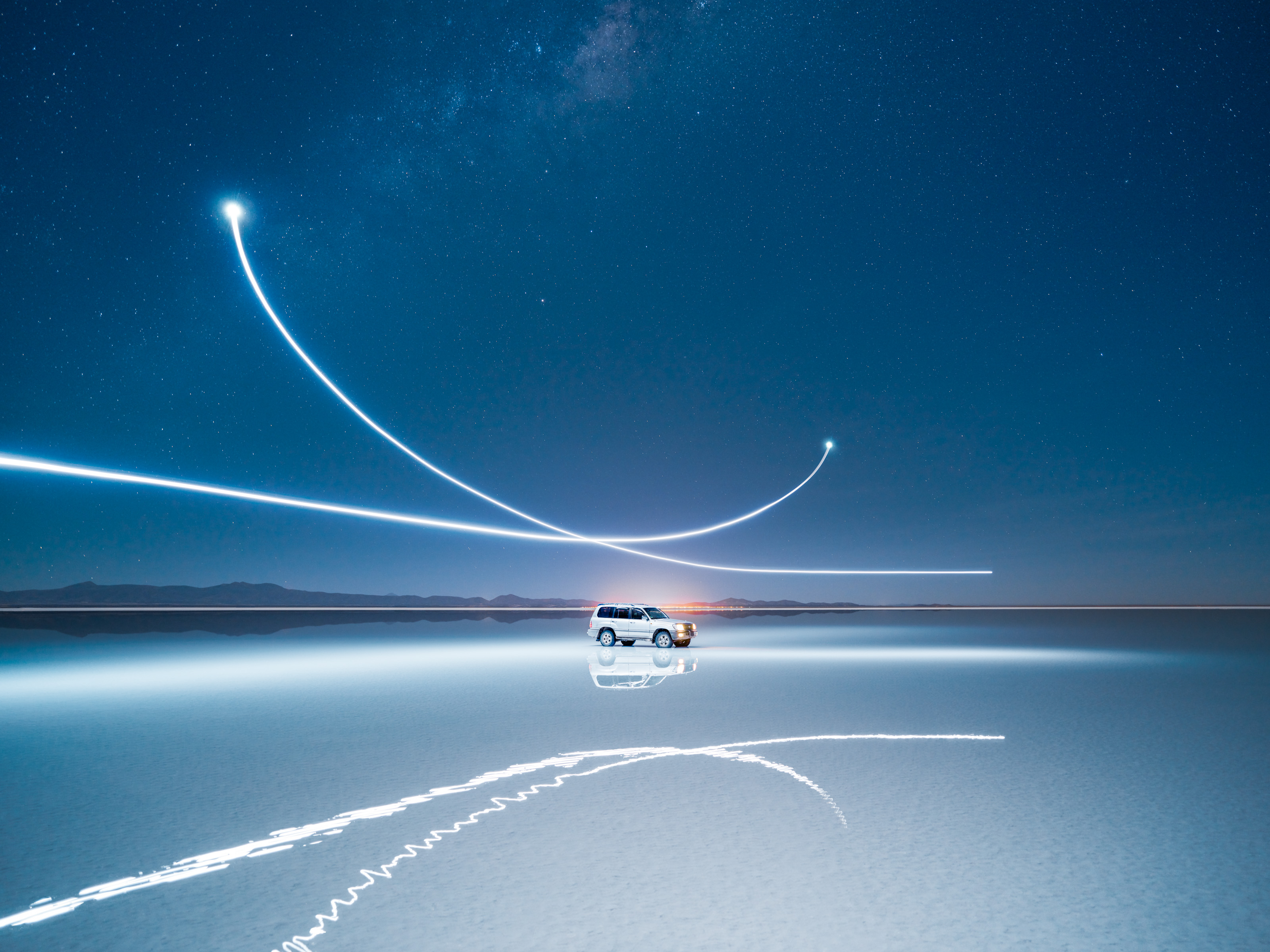 A car on a reflective surface under a starry sky, surrounded by mesmerizing drone light painting trails—capturing the magic of nighttime landscape photography.