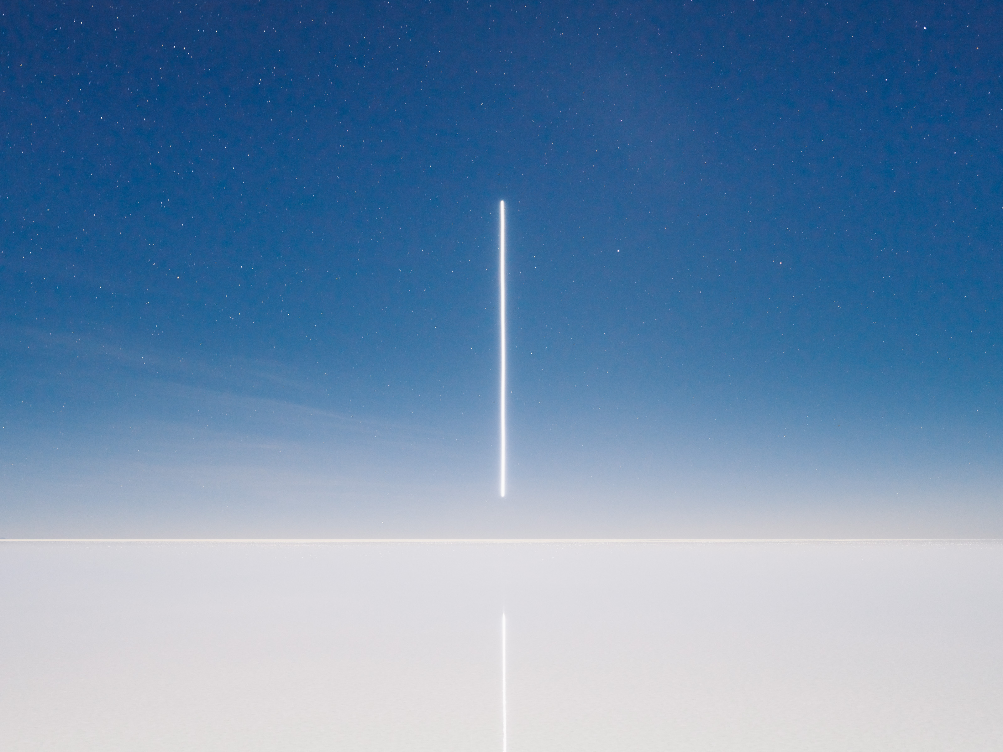 A vertical white streak of drone light art extends from the horizon, reflected on a calm, flat surface under a starry night sky.