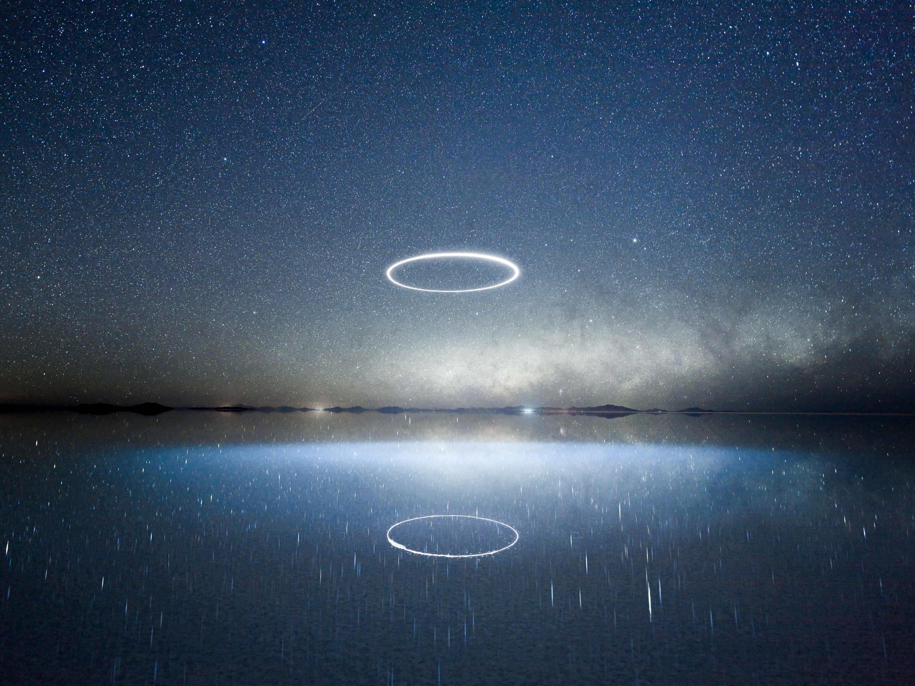 A glowing, futuristic ring hovers in a starry night sky, reflected in a calm, mirror-like body of water—a striking scene reminiscent of nighttime landscape photography.