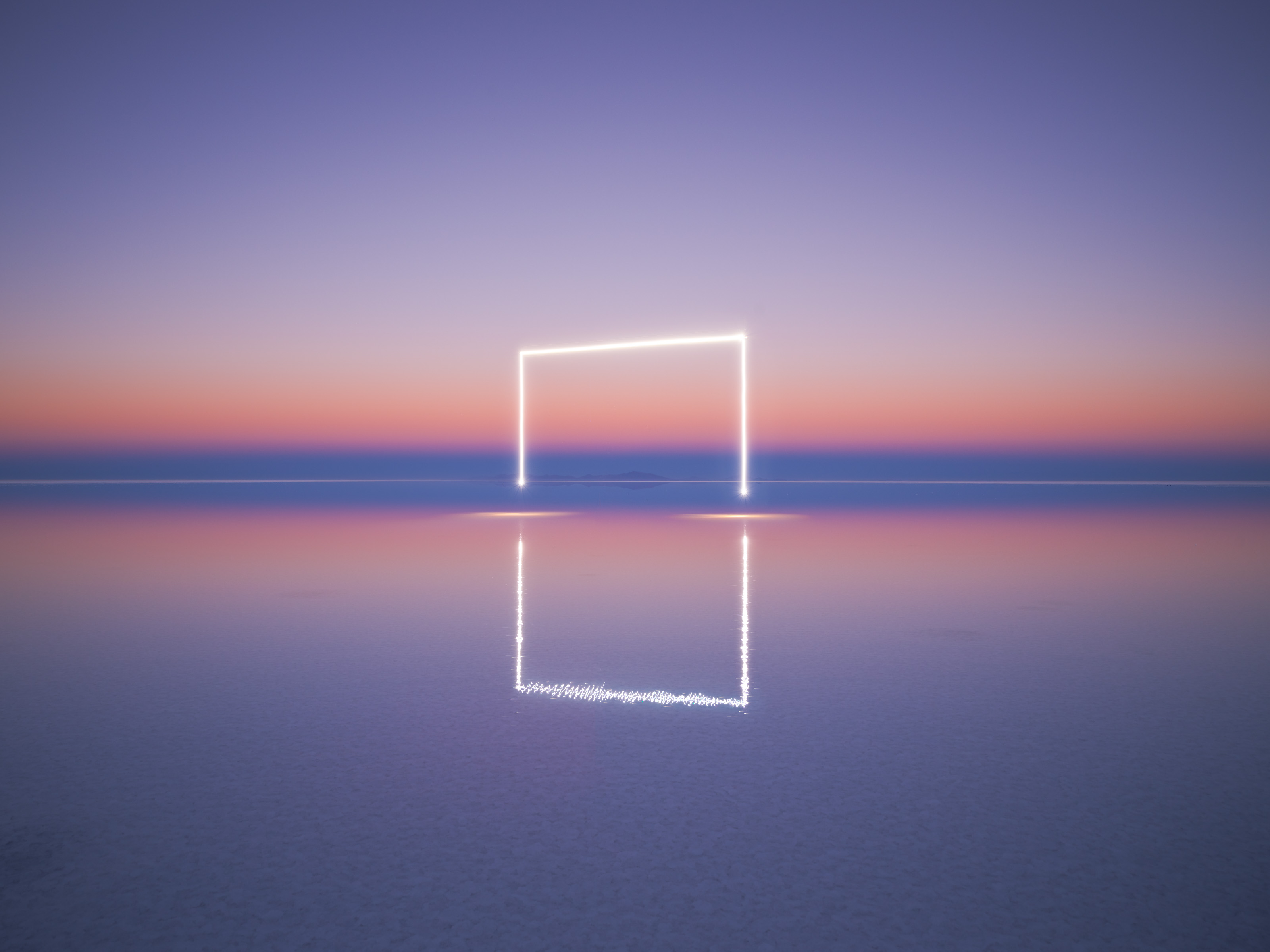 A glowing square frame, created by drone light painting, stands above calm water, reflecting a pastel sunset sky.