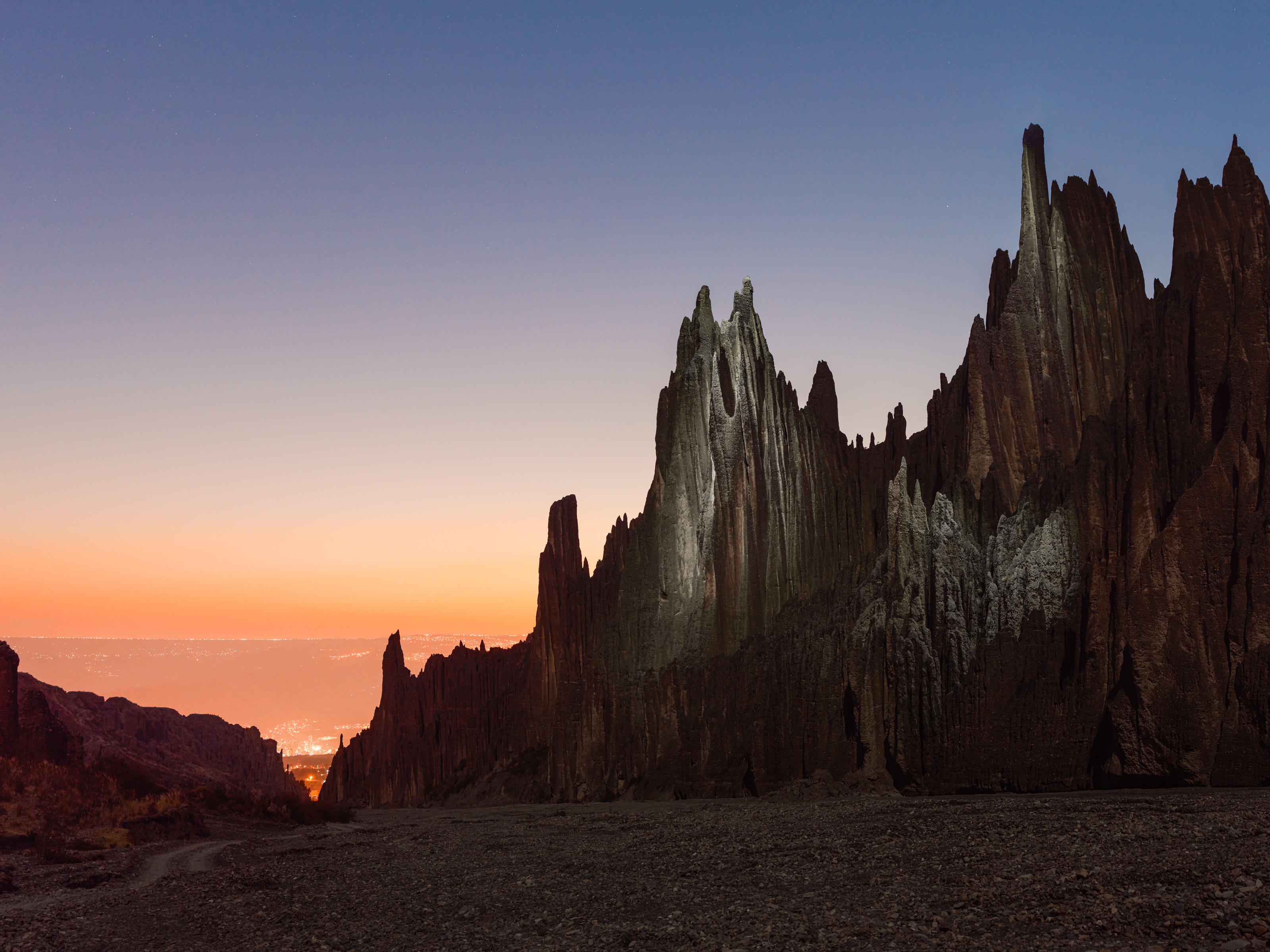 Jagged rock formations at dusk with city lights glowing in the distance under a clear sky, captured by a photographer experimenting with drone light painting.