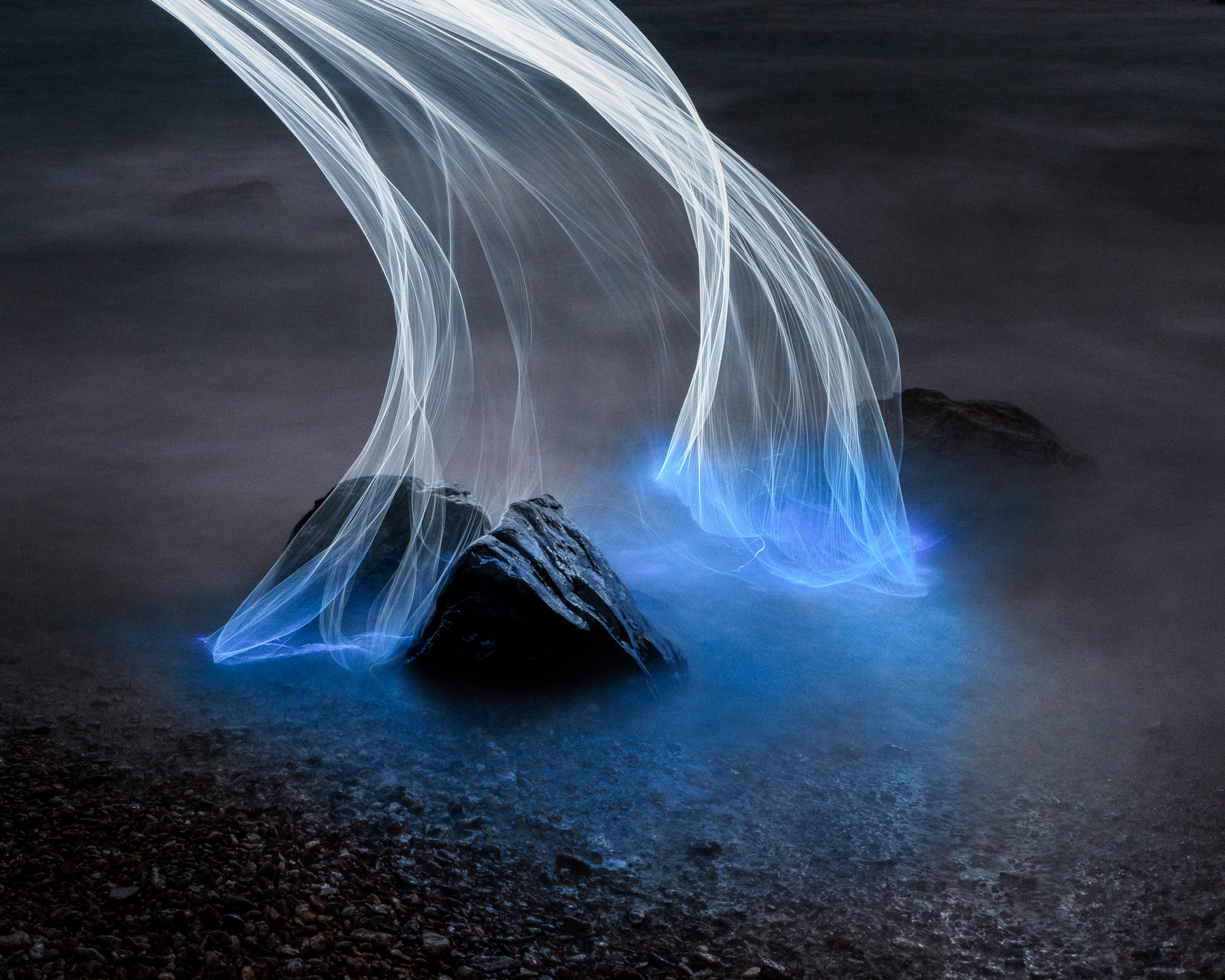 Long-exposure photo of rocks with flowing blue and white light trails above misty water.
