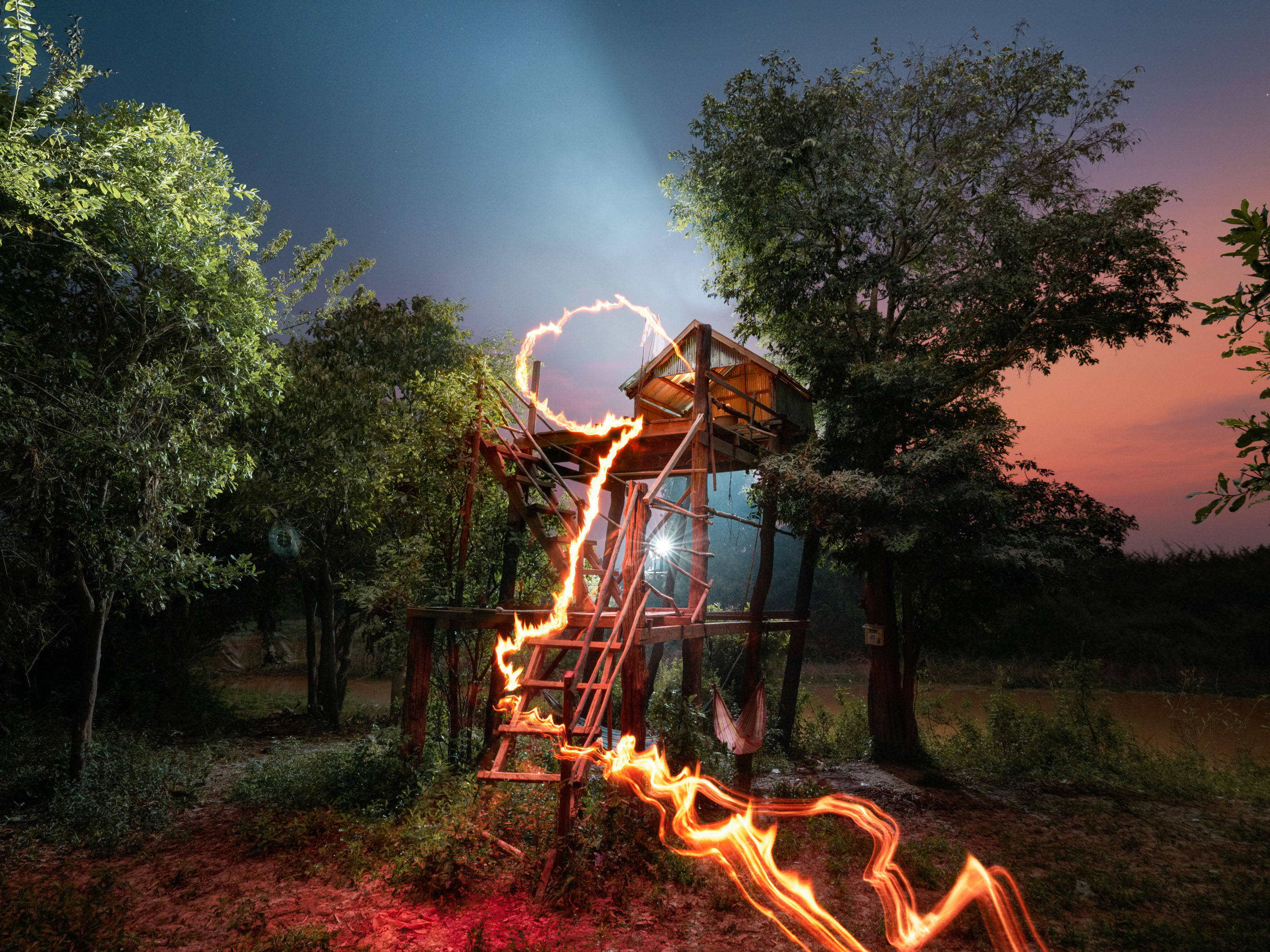 A glowing drone light trail leads to a treehouse at dusk, surrounded by trees under a colorful twilight sky, creating a touch of futuristic magic.