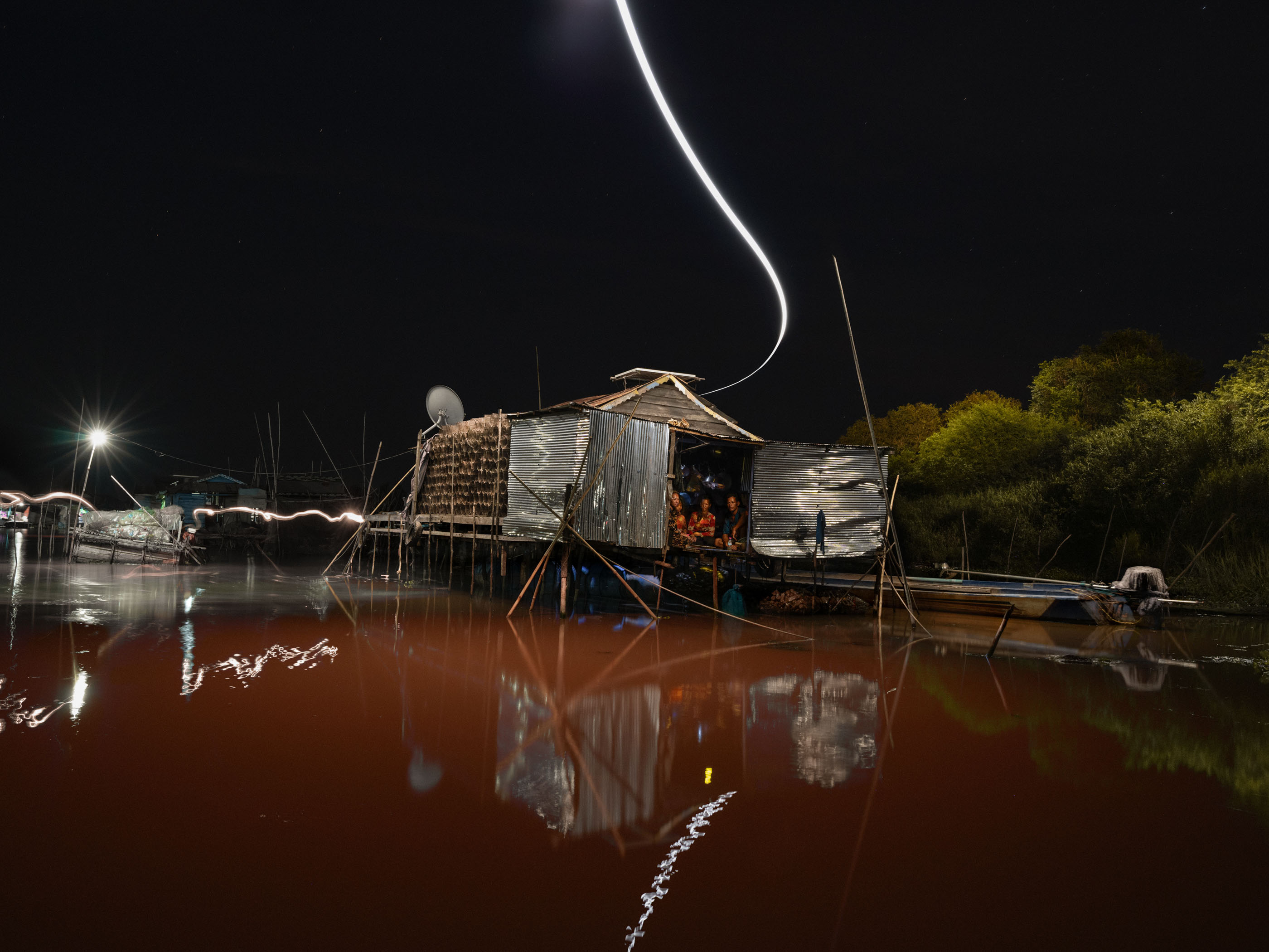A rustic riverside shack at night becomes a futuristic scene, as a bright light trail curves above and reflects on red water—perfect for nighttime landscape photography.
