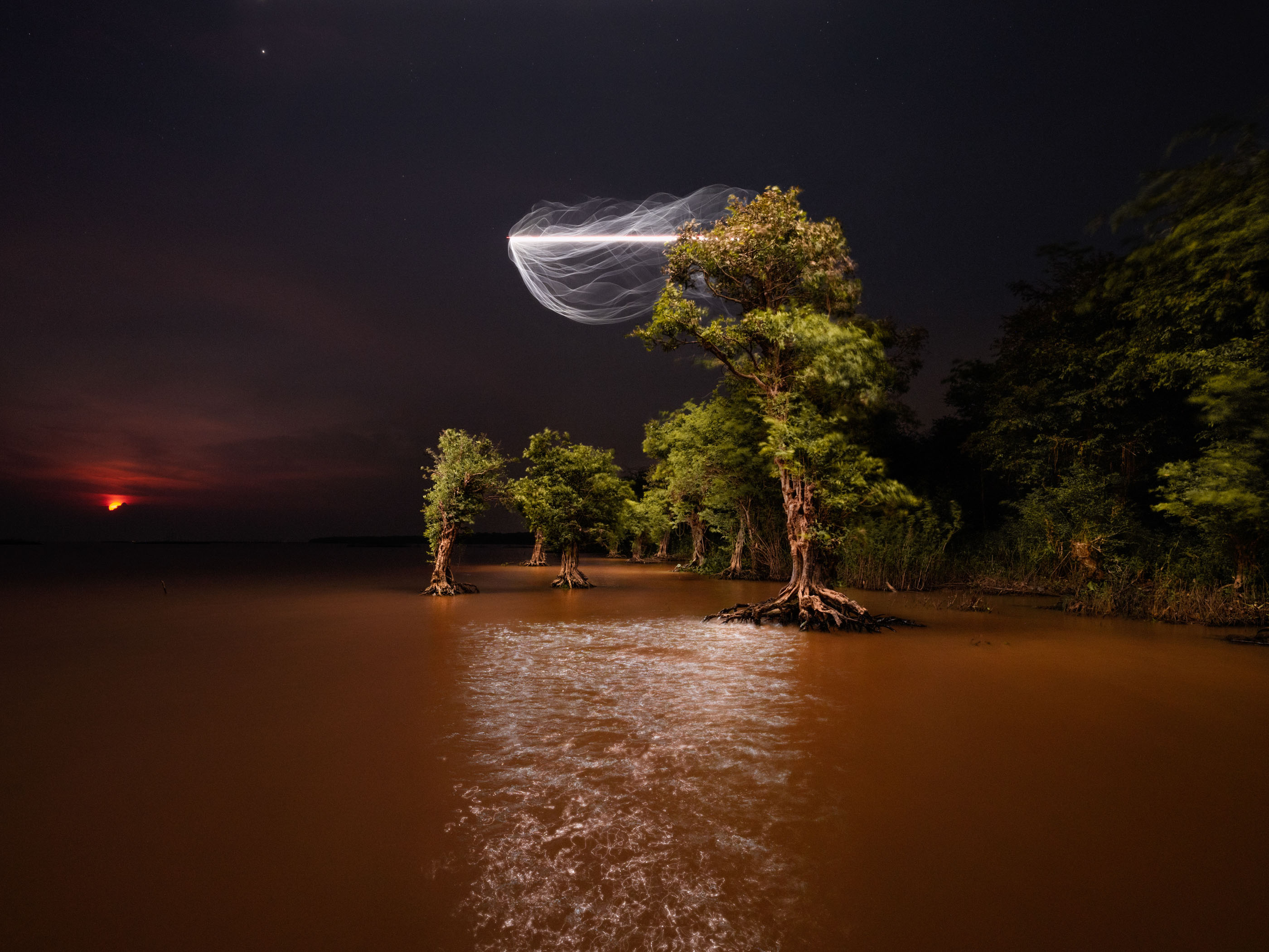 Night scene with trees in water, a red moon, and a glowing, swirling drone light art display illuminating the sky above the trees.
