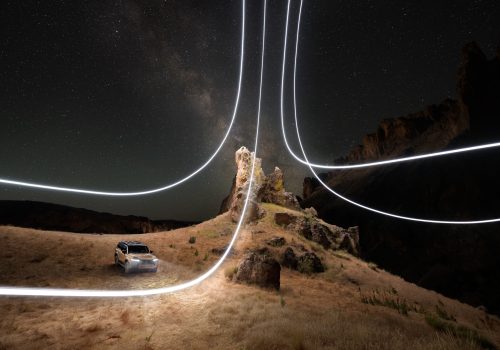 A car on dry grass at night with glowing light trails and a starry sky in the background, captured by a photographer skilled in nighttime landscape photography.