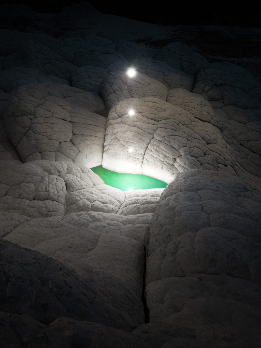A green pool surrounded by rocky white terrain glows under three bright lights above it at night, capturing the magic of nighttime landscape photography.