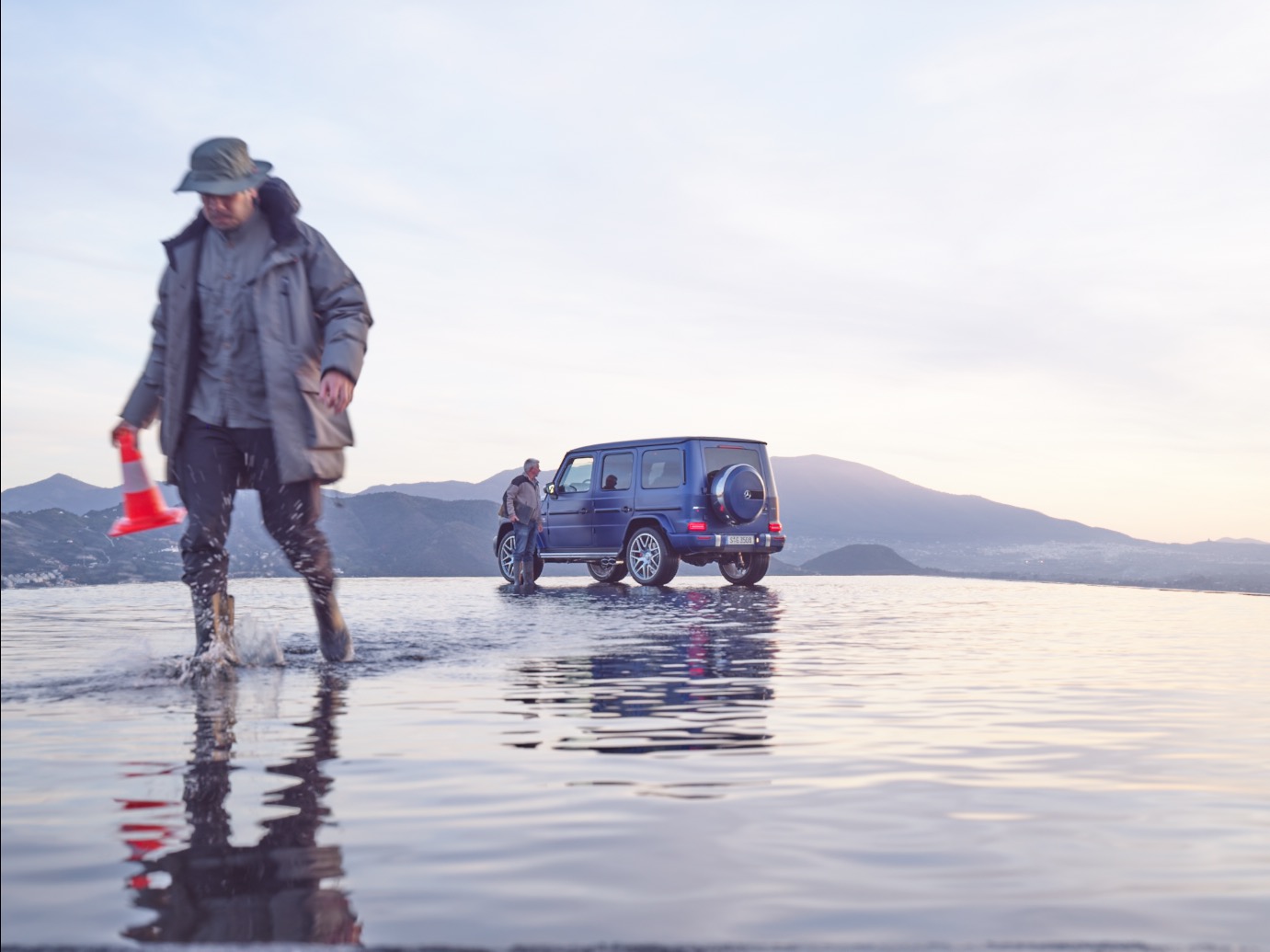 Two people stand near a blue SUV parked in shallow water, with mountains in the background, creating a futuristic scene reminiscent of nighttime landscape photography.