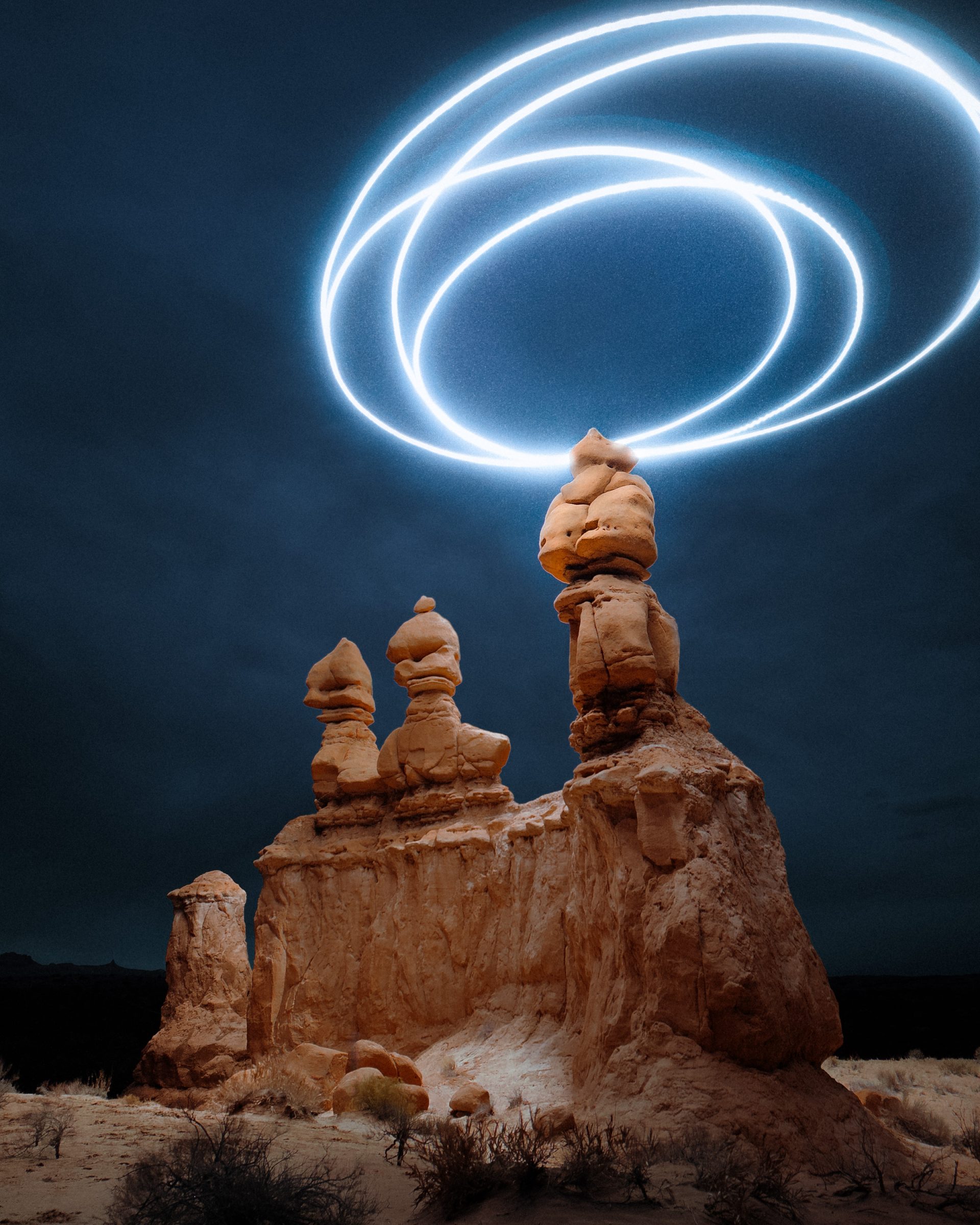 Three tall rock formations at night with futuristic glowing light trails swirling in the sky above them, created by a photographer using drone light painting techniques.