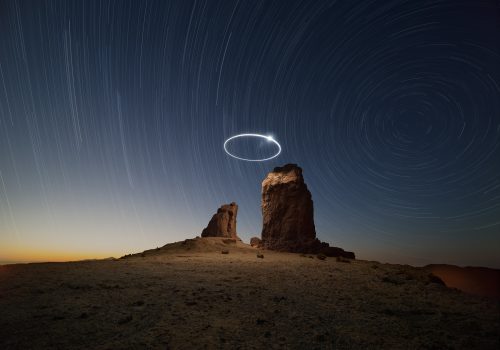 Two large rock formations at night, captured by a fine artist, stand beneath swirling star trails and a bright circular drone light art display in the sky.