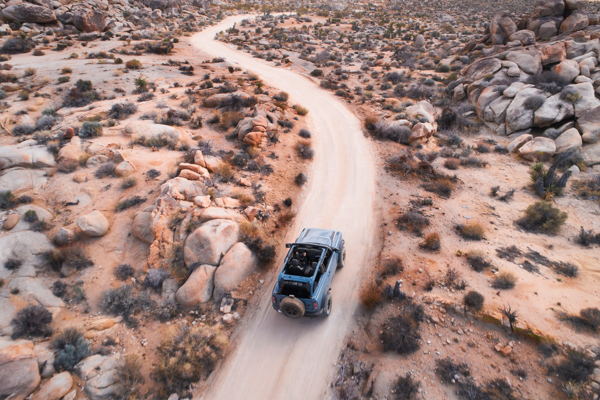 A blue SUV drives on a winding dirt road through a rocky, desert landscape with sparse shrubs, as a photographer captures the adventure.
