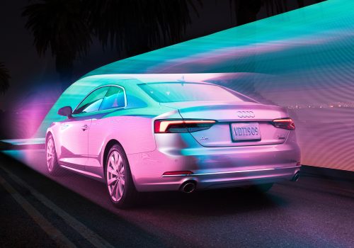 A silver Audi car drives at night with colorful neon light trails surrounding it, creating a futuristic scene reminiscent of drone light painting.
