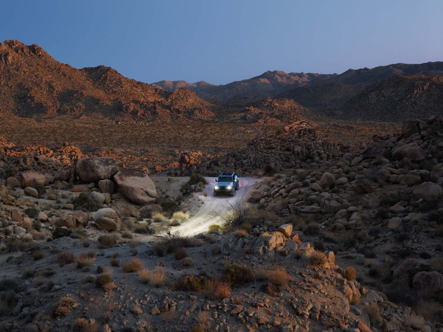 A car with headlights on drives through a rocky desert landscape at dusk, surrounded by mountains—perfect for nighttime landscape photography.