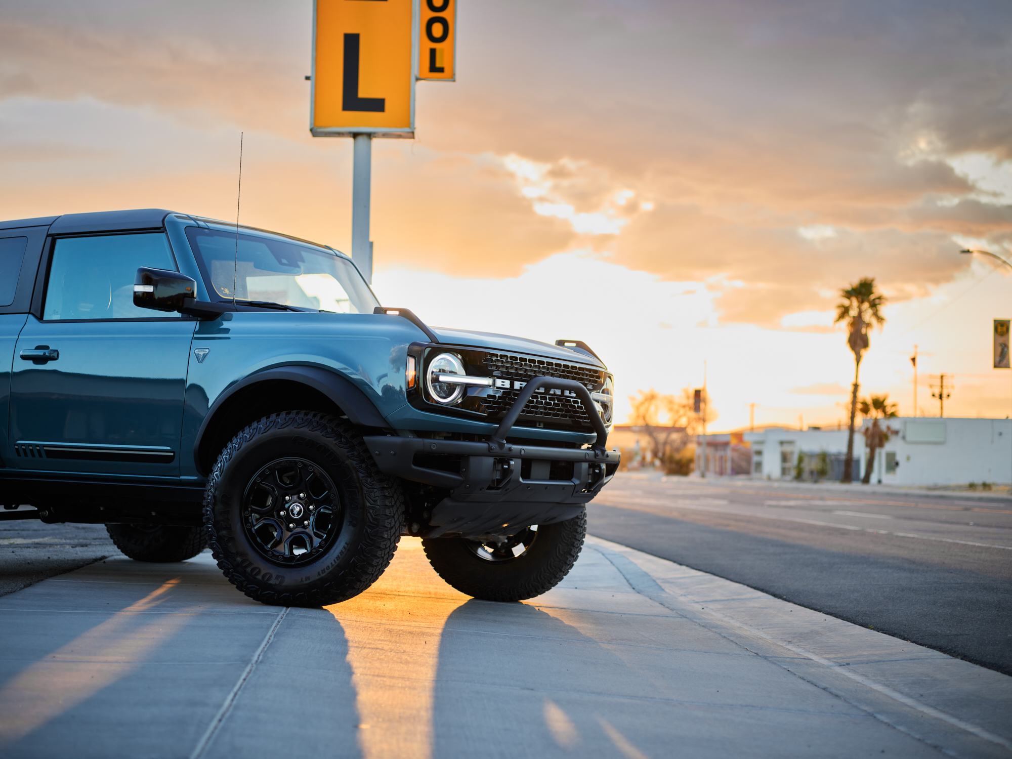 Blue Ford Bronco parked on a city street at sunset, with a yellow sign and palm trees in the background, illuminated by futuristic drone light art that adds a creative glow to the scene.