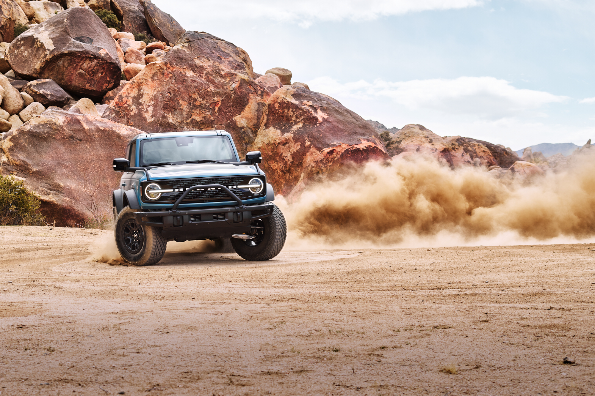 A blue Ford Bronco kicks up dust while driving off-road near large boulders, as a photographer captures the desert landscape, illuminated by creative drone light painting.