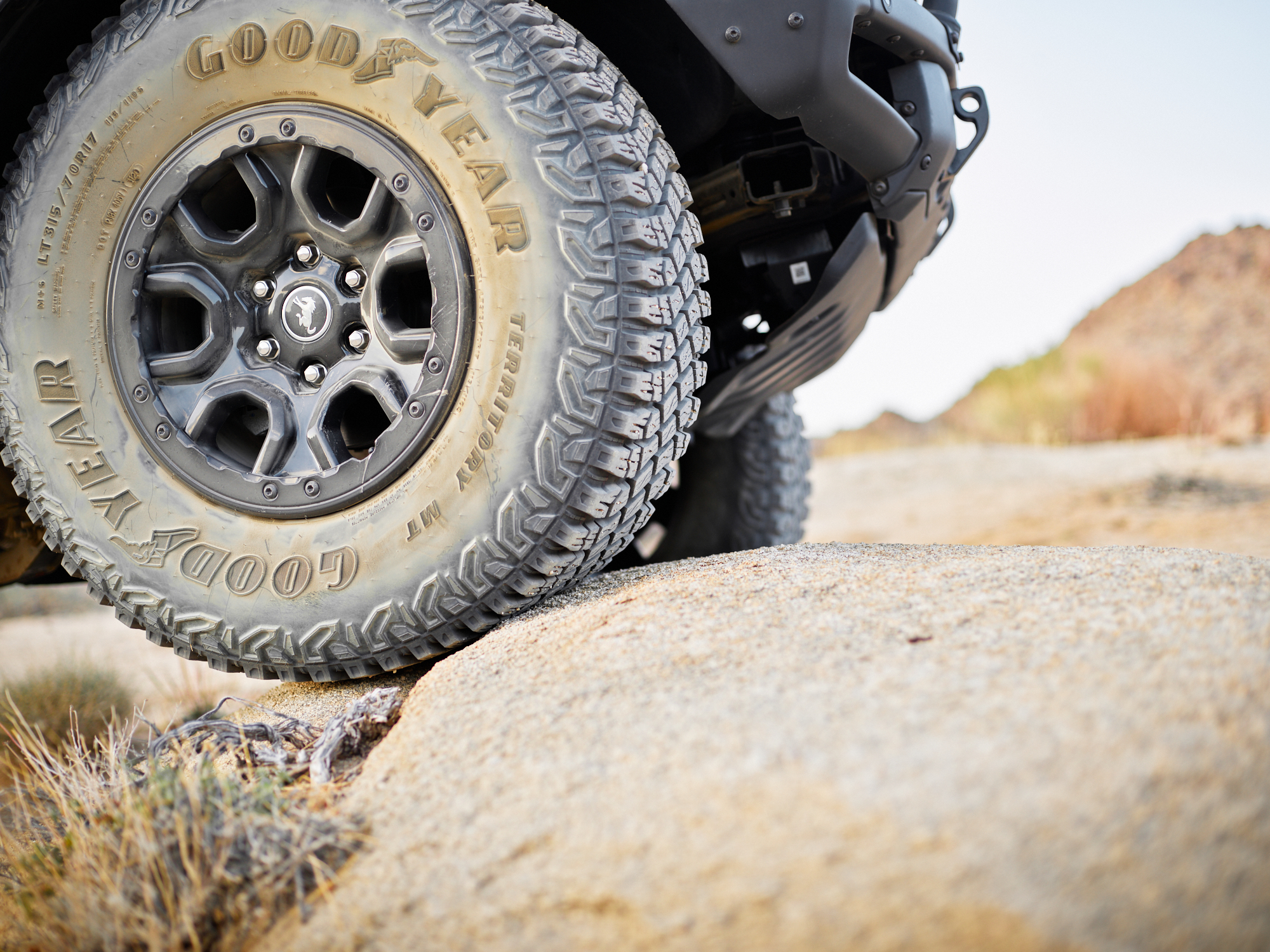 Close-up of a rugged Goodyear tire on a vehicle driving over a large rock in a rocky, desert landscape, with drone light painting subtly illuminating the scene like the touch of a fine artist.
