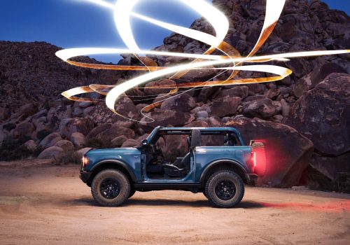 A teal off-road SUV parked on sand at dusk, surrounded by swirling drone light art, with rocks in the background—a striking example of futuristic nighttime landscape photography.