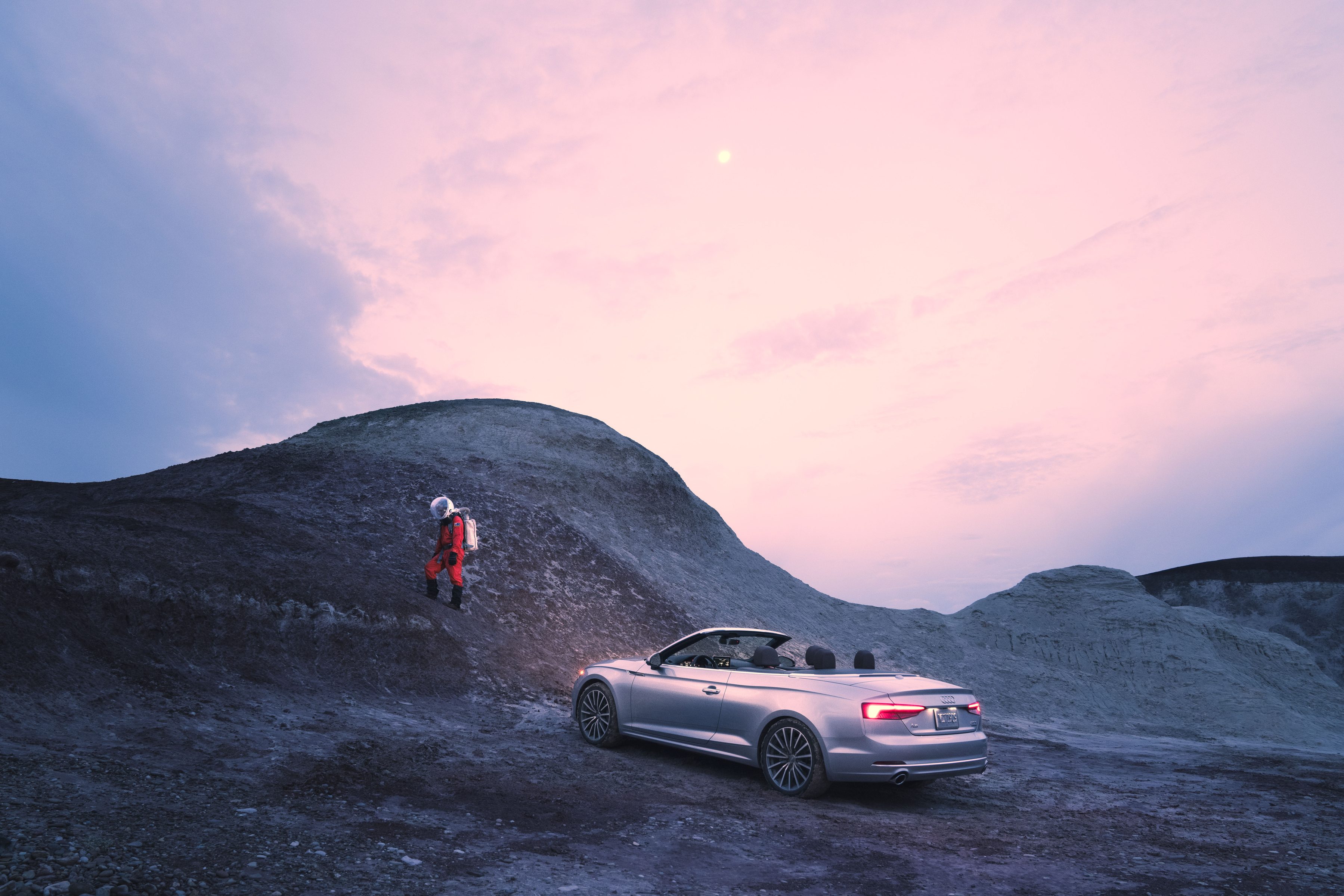 A futuristic scene unfolds as a person with a jetpack soars near a silver convertible on rocky terrain at sunset.